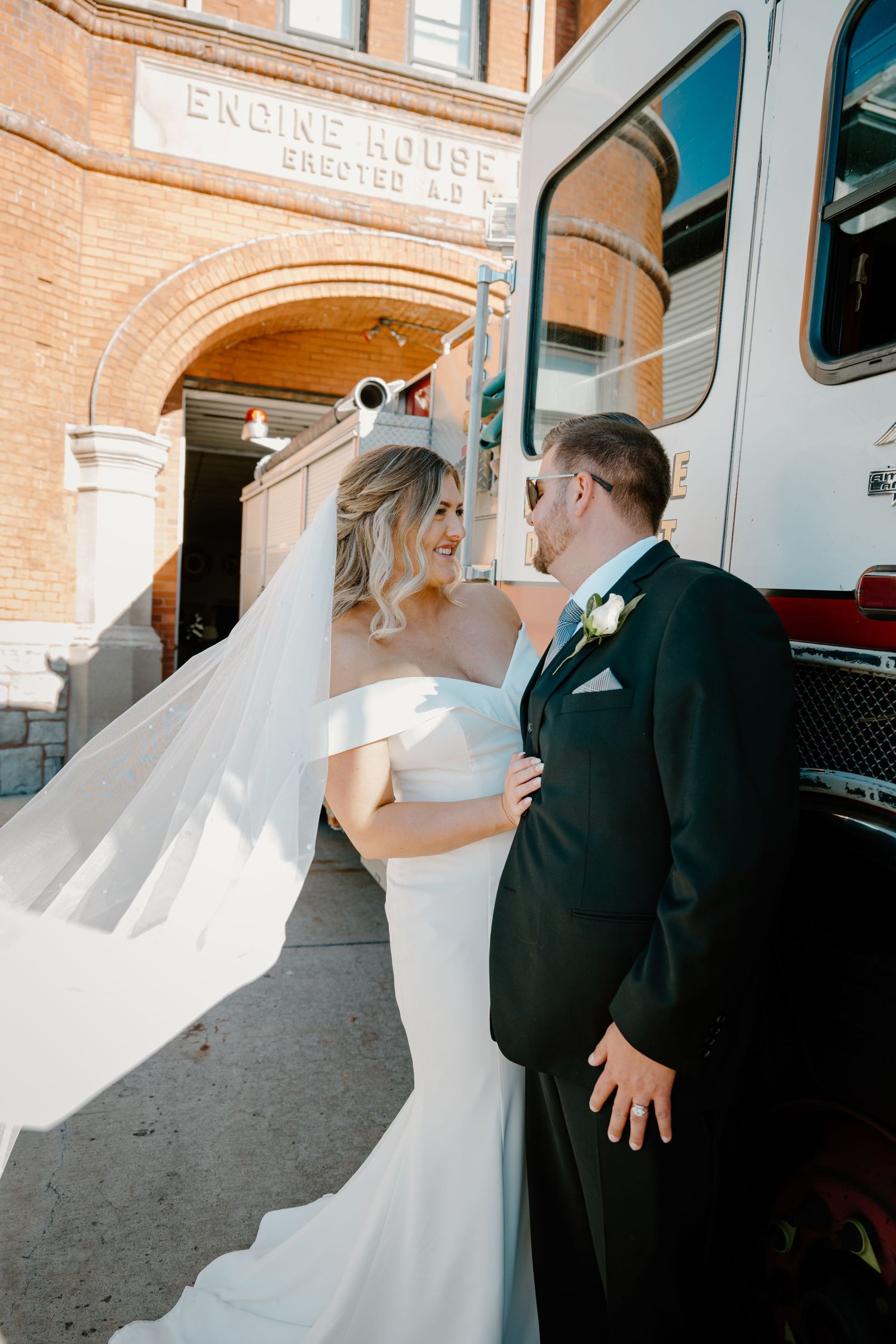 Bride in flowing veil and groom share tender moment beside fire truck at historic engine house.