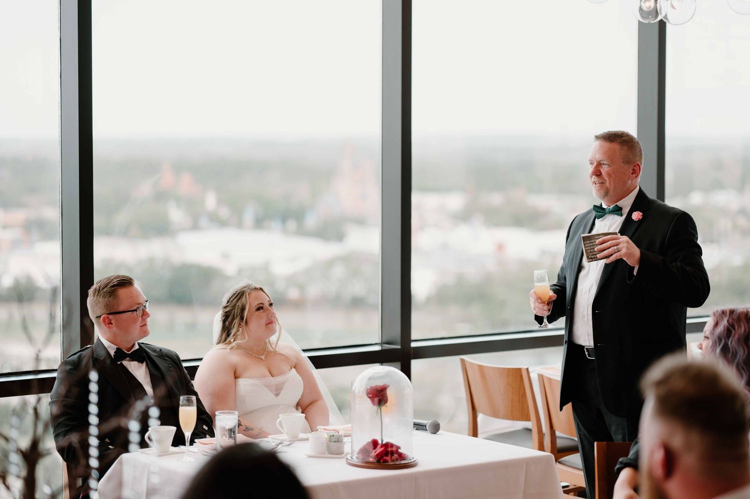 Groom giving a toast speech to bride and groom seated at wedding reception table with panoramic windows.