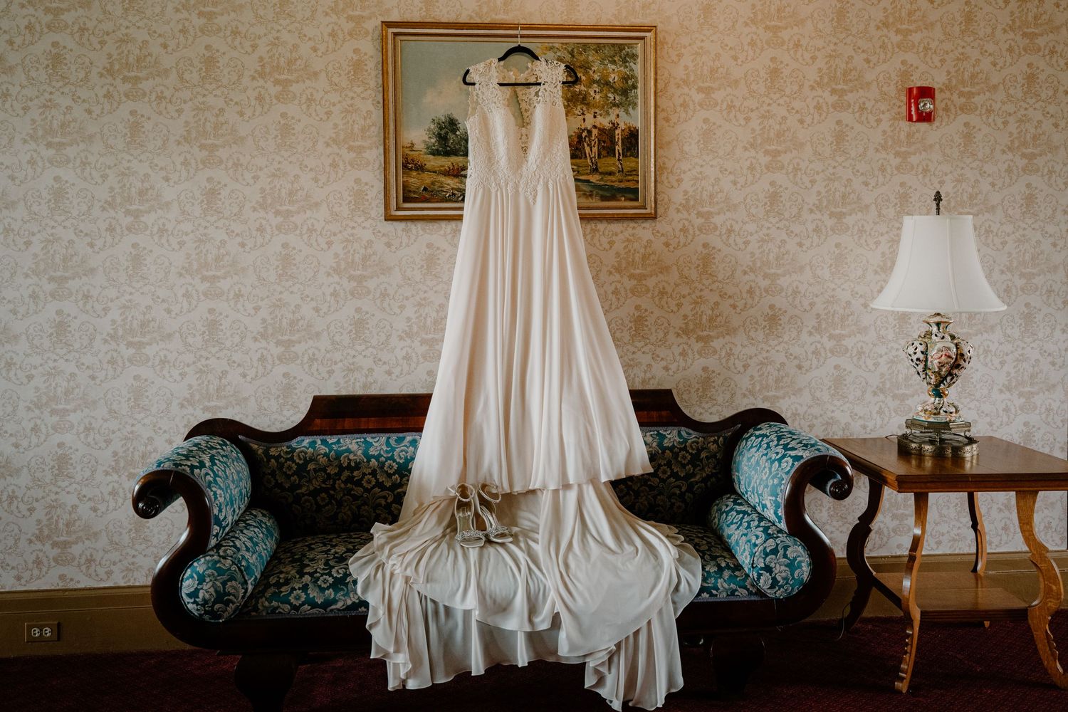 Simple white A-line wedding dress displayed on hanger above antique teal sofa in vintage wallpapered room.