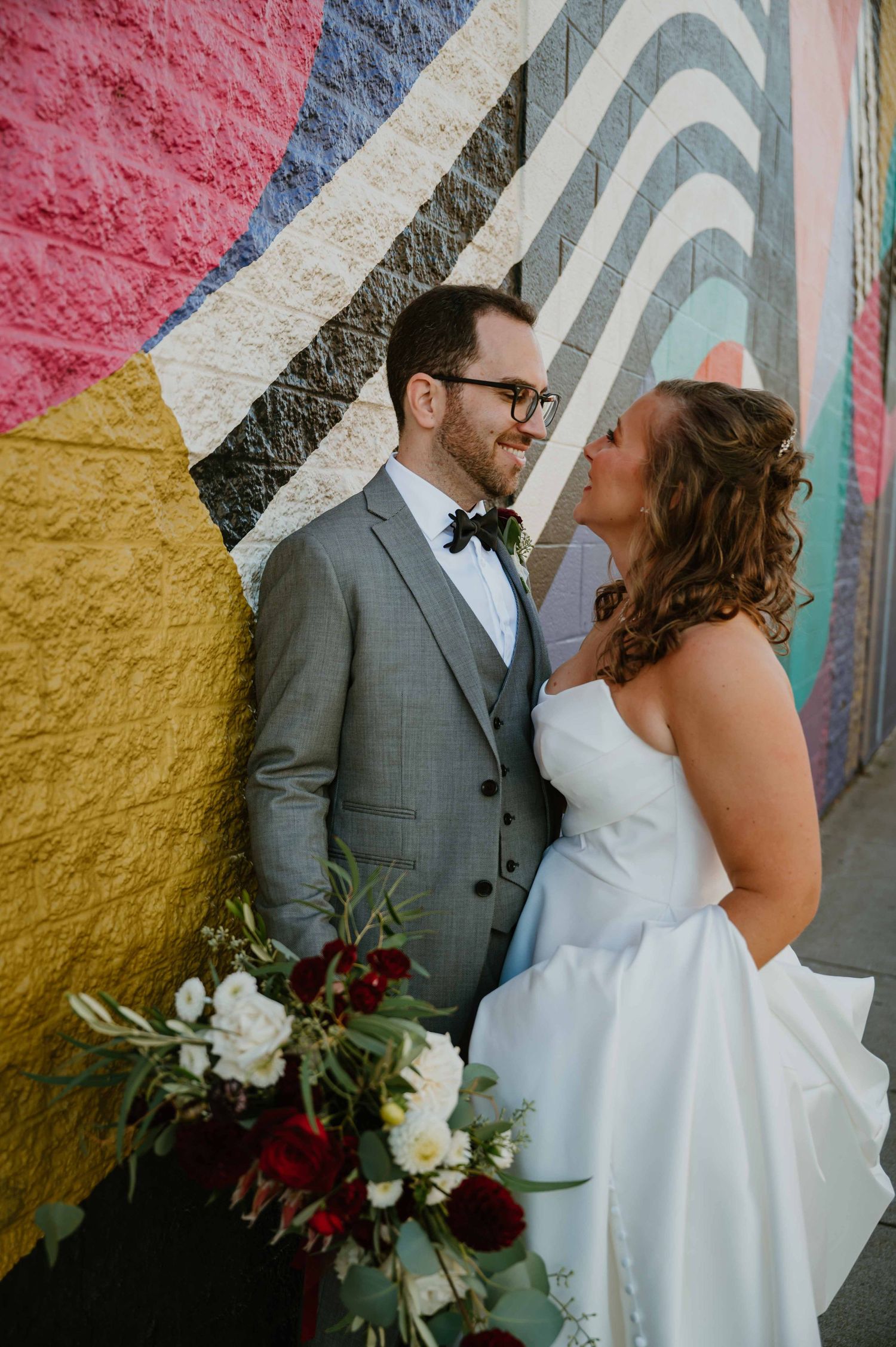 Bride and groom smiling at each other against vibrant colorful mural holding bridal bouquet.
