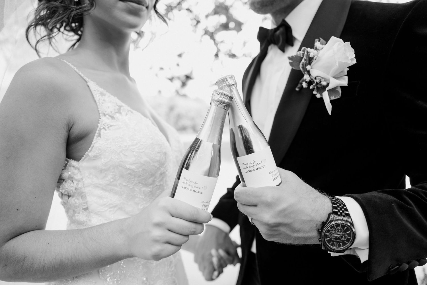 Bride and groom clinking champagne bottles in black and white wedding photo.