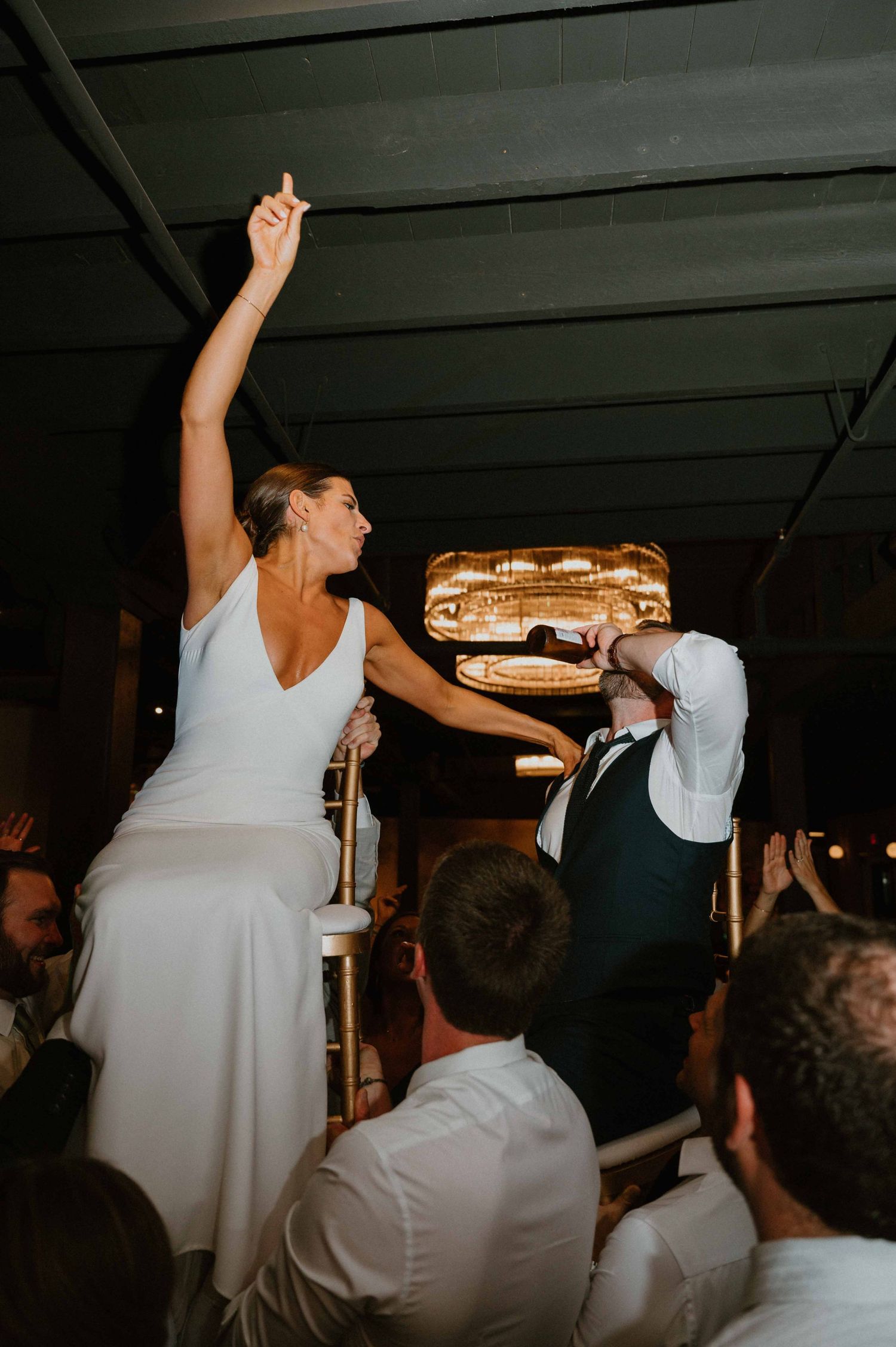 Bride raises arm triumphantly while being lifted by guests during lively wedding reception celebration.