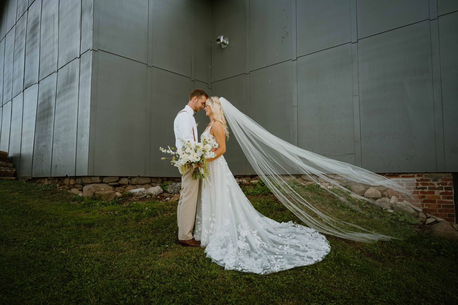 Bride and groom sharing romantic kiss with flowing cathedral veil against modern gray barn wall.