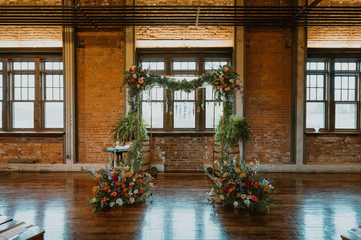 Colorful floral wedding arch with greenery stands in a rustic brick industrial venue with wooden floors.