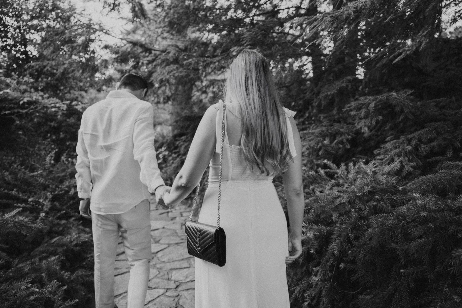 Black and white photo of couple walking hand in hand along garden path surrounded by lush greenery.