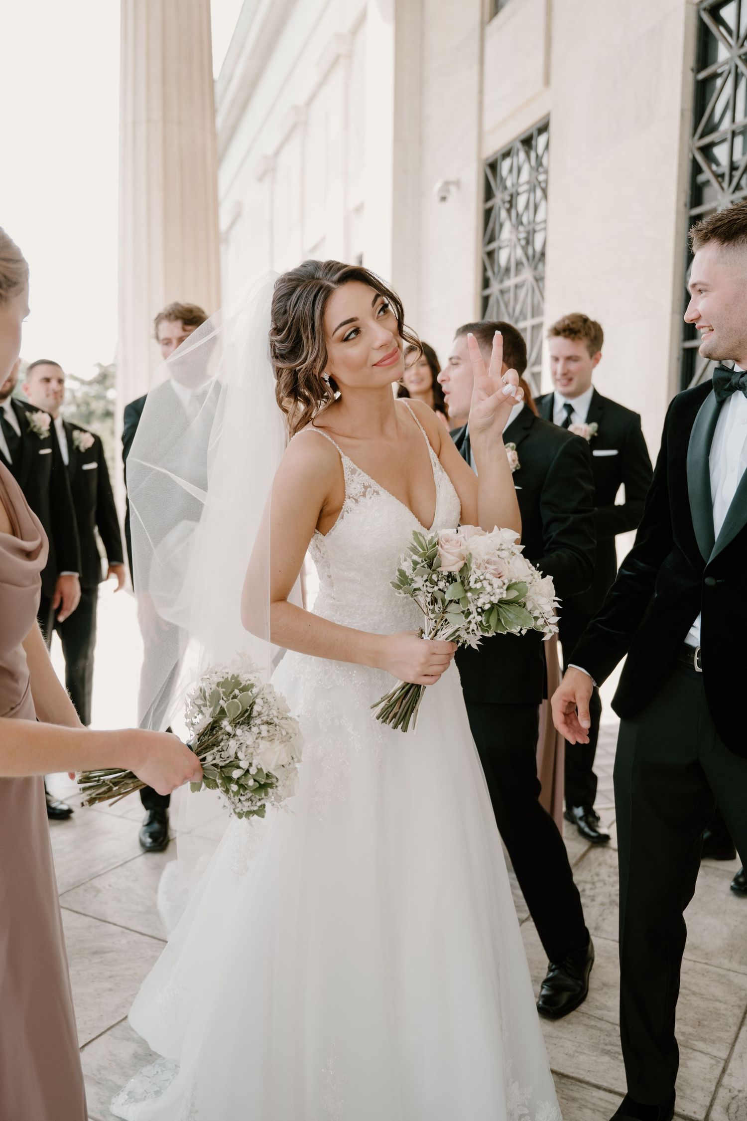 Smiling bride in lace wedding gown holding bouquet surrounded by wedding party outdoors.