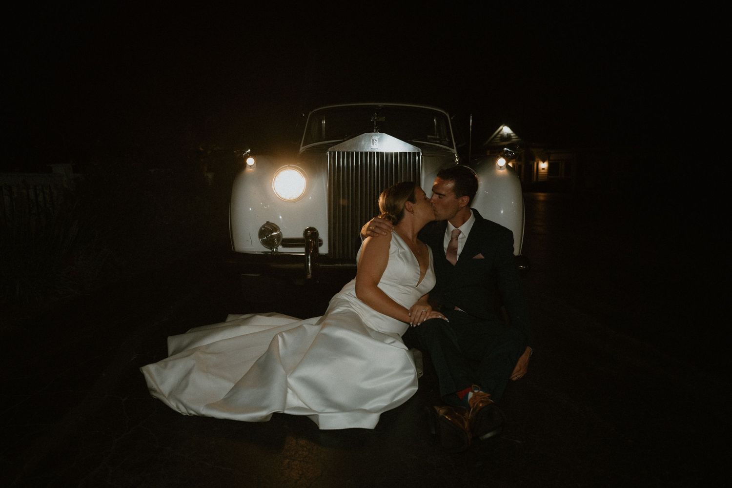 Bride and groom kissing in front of a vintage white Rolls Royce at night with soft lighting.