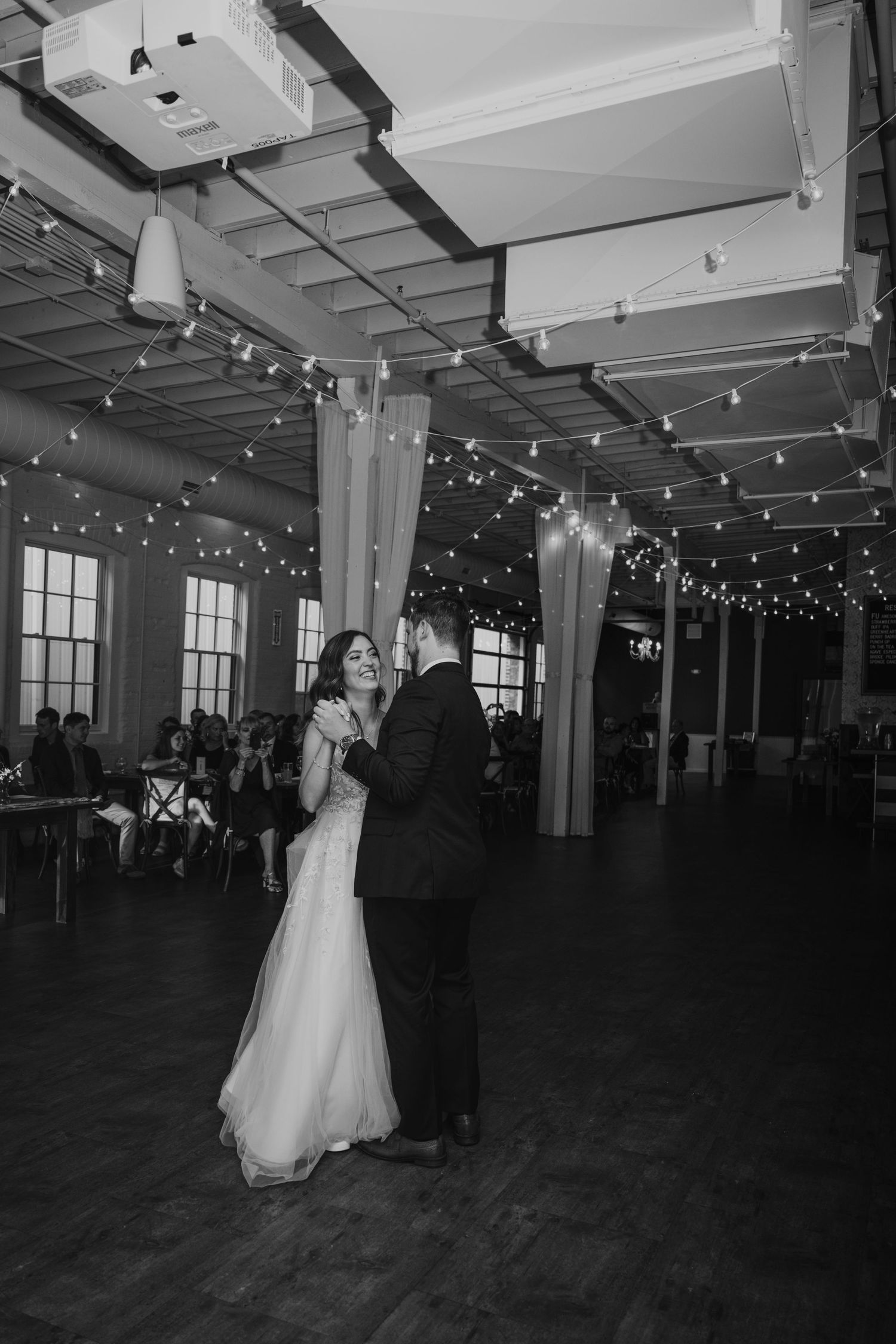 Bride and groom share first dance under string lights in elegant industrial wedding venue.