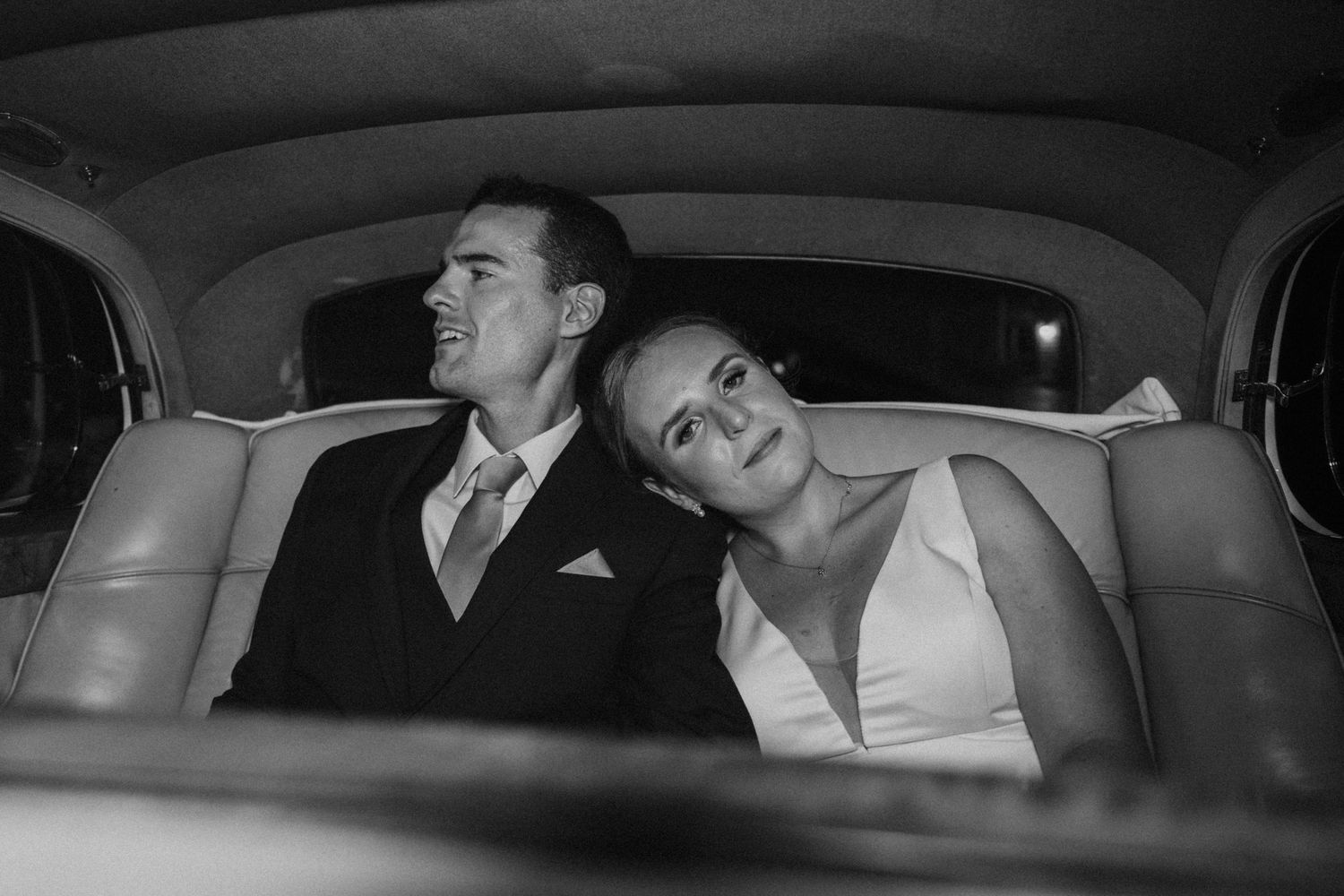 Black and white photo of bride and groom sitting together in the back seat of a classic car.