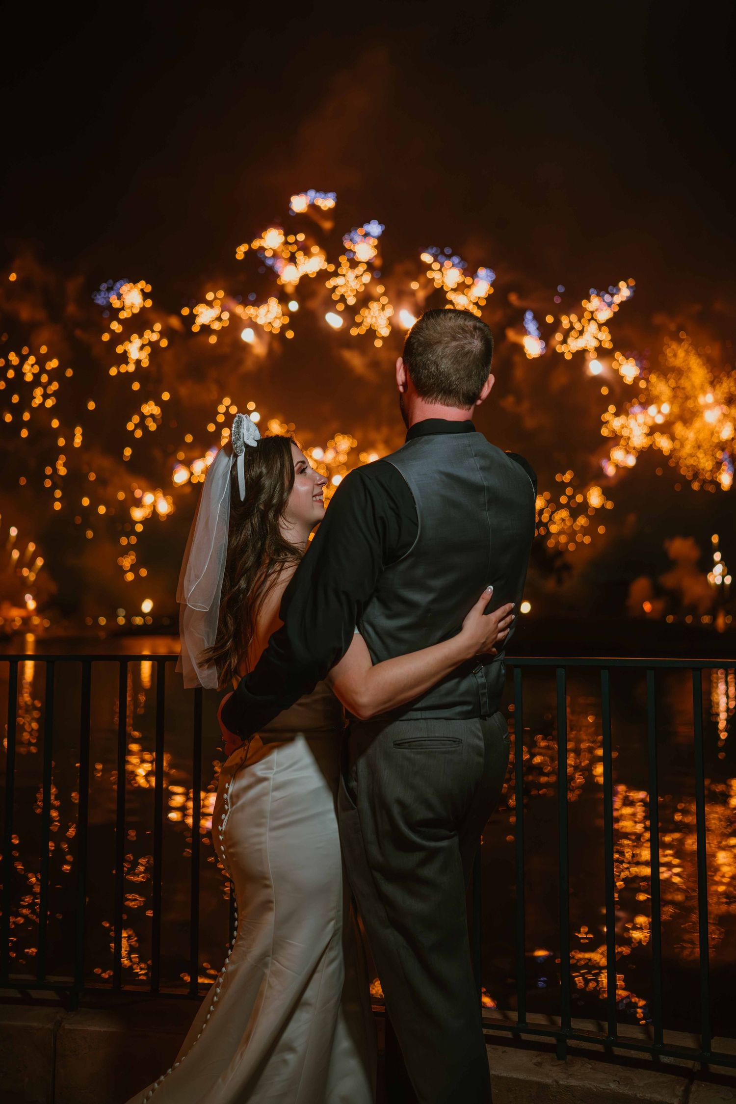 Newlyweds embrace on balcony watching brilliant fireworks burst over water on their wedding night.