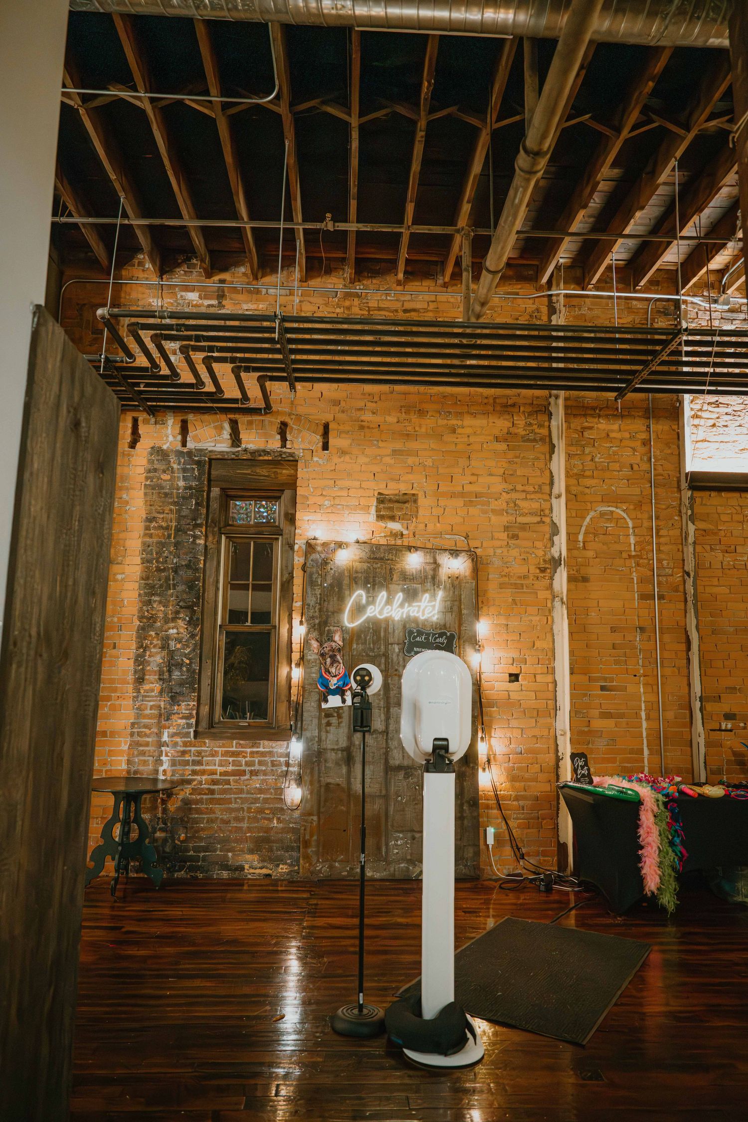 Rustic brick wall wedding photo booth with neon celebrate sign and string lights.