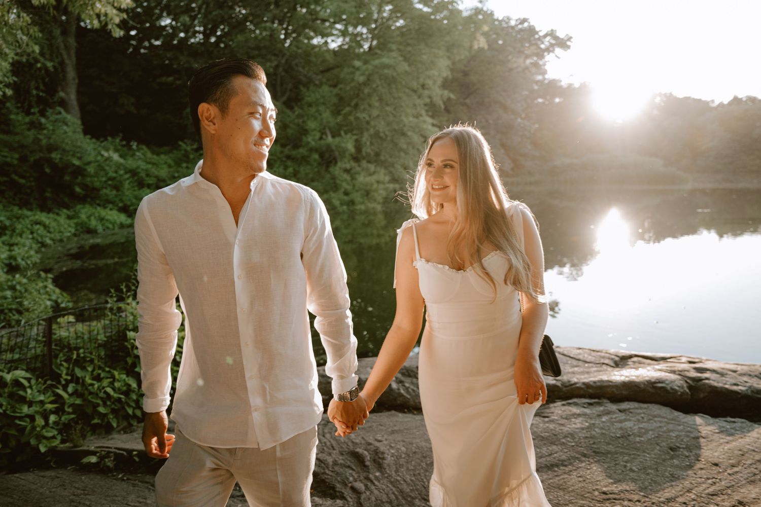 Happy couple walks hand in hand near sunlit lakeside, smiling at each other during golden hour.