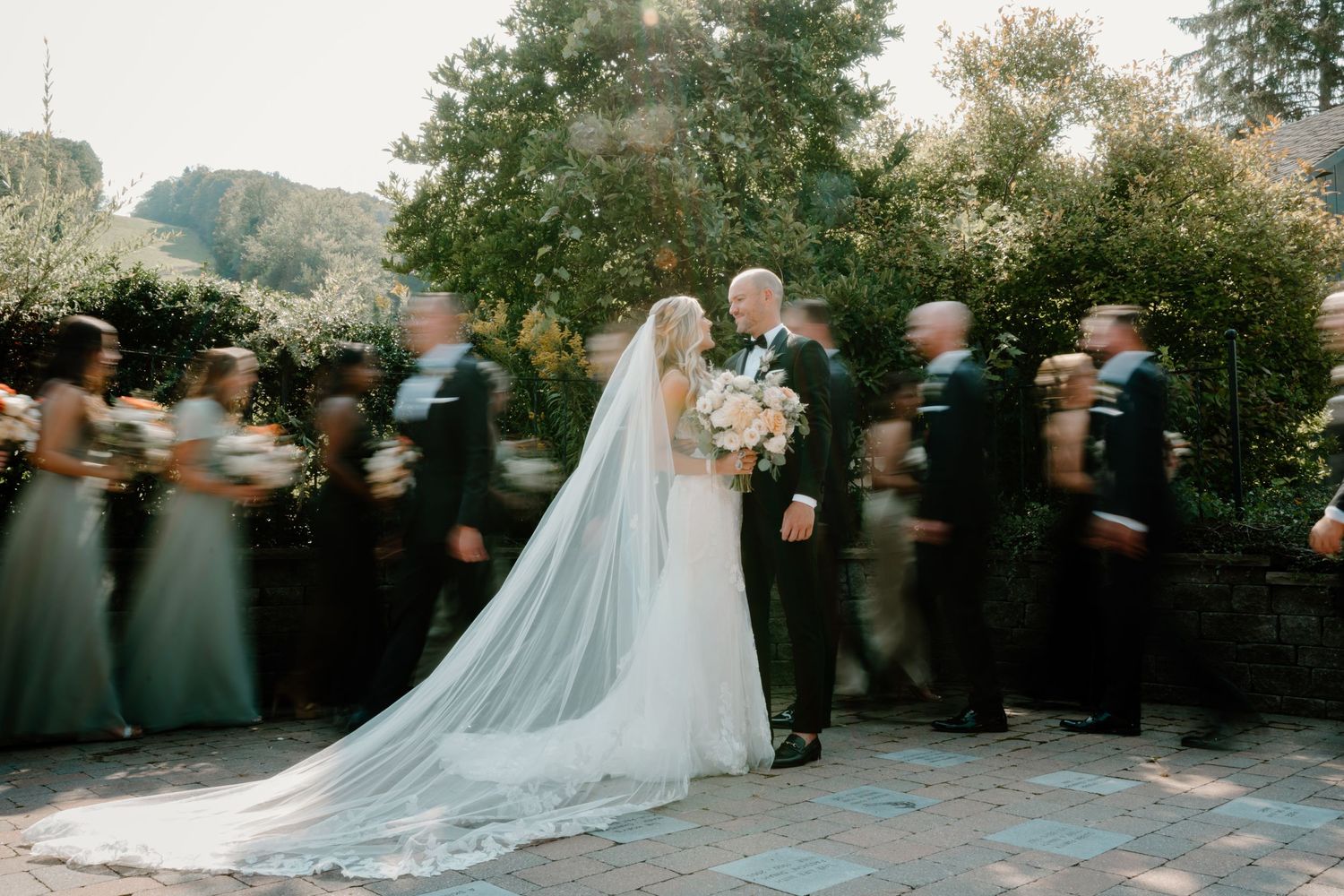 Wedding party in motion blur surrounds bride and groom in garden setting.