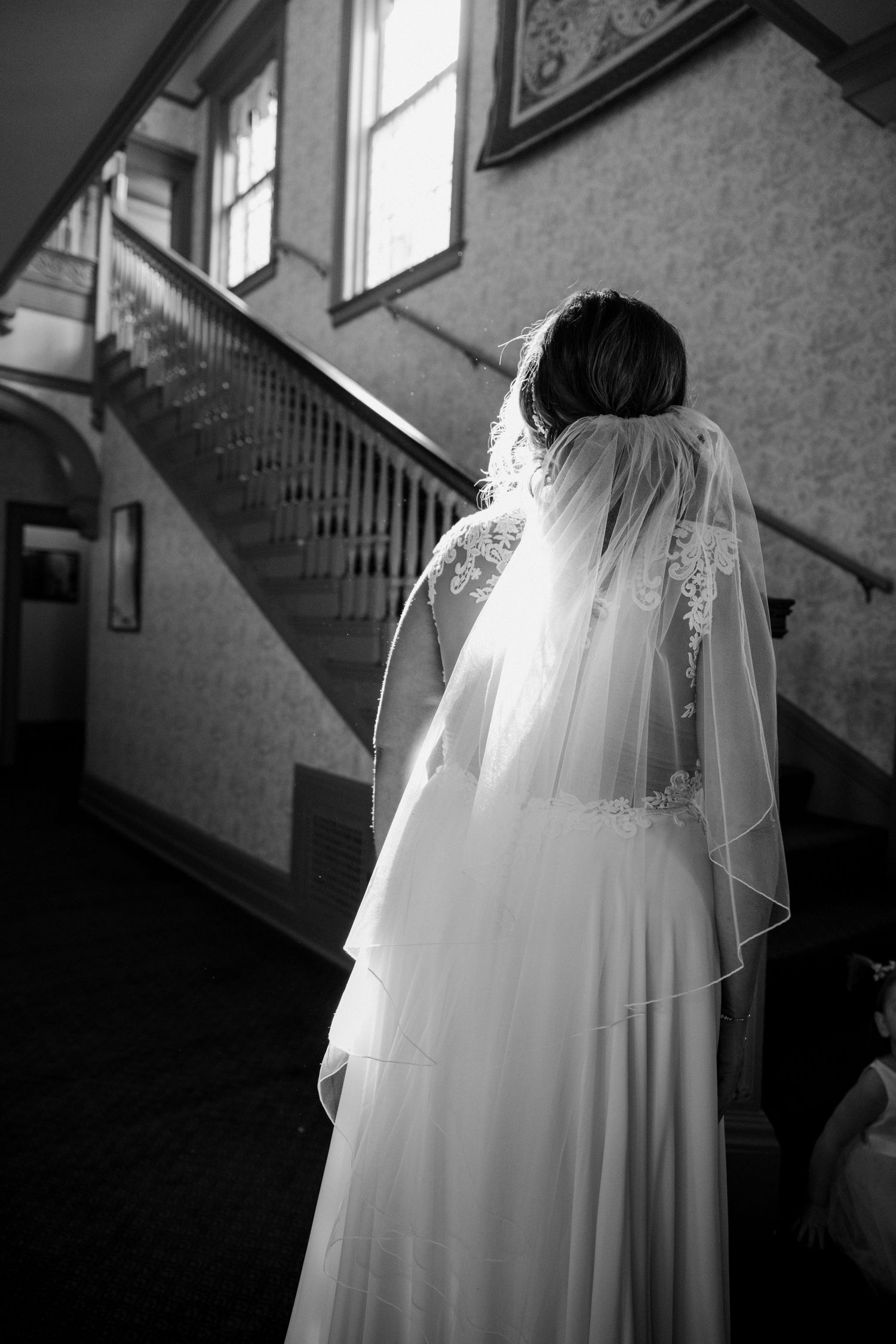 Black and white photo of a bride in a lace-trimmed veil walking toward a grand staircase.