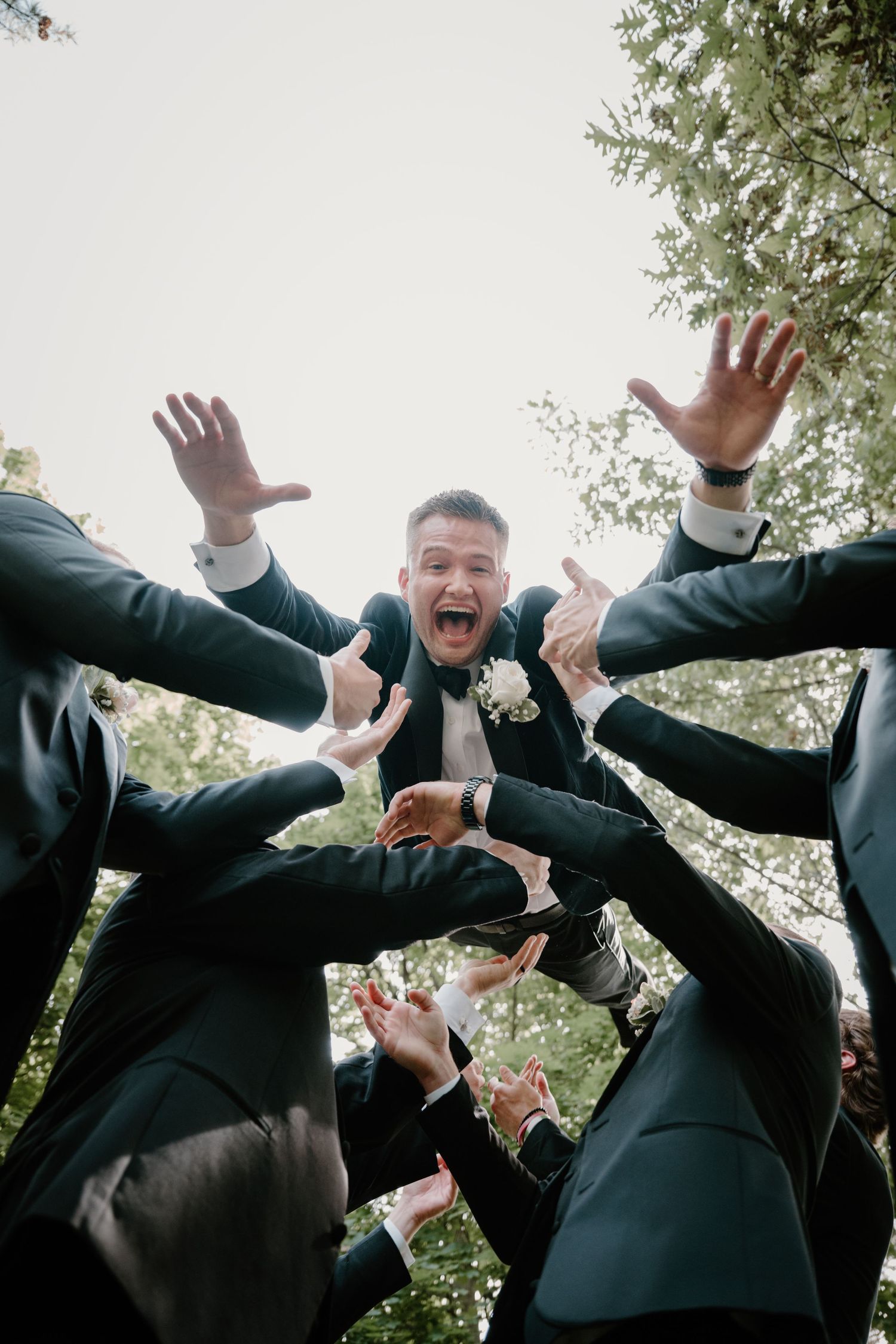 Excited groom being tossed by groomsmen in dark suits outdoors at wedding celebration.