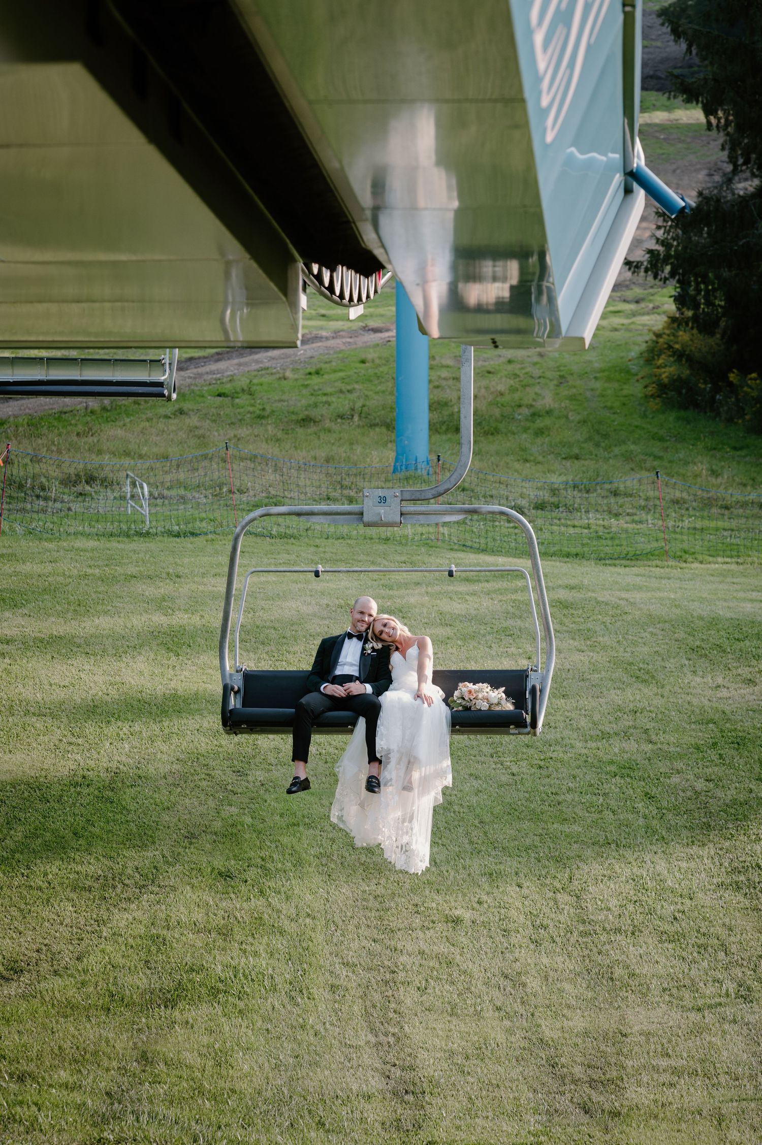 Bride and groom sharing a romantic kiss on a ski lift chair above a green summer slope.