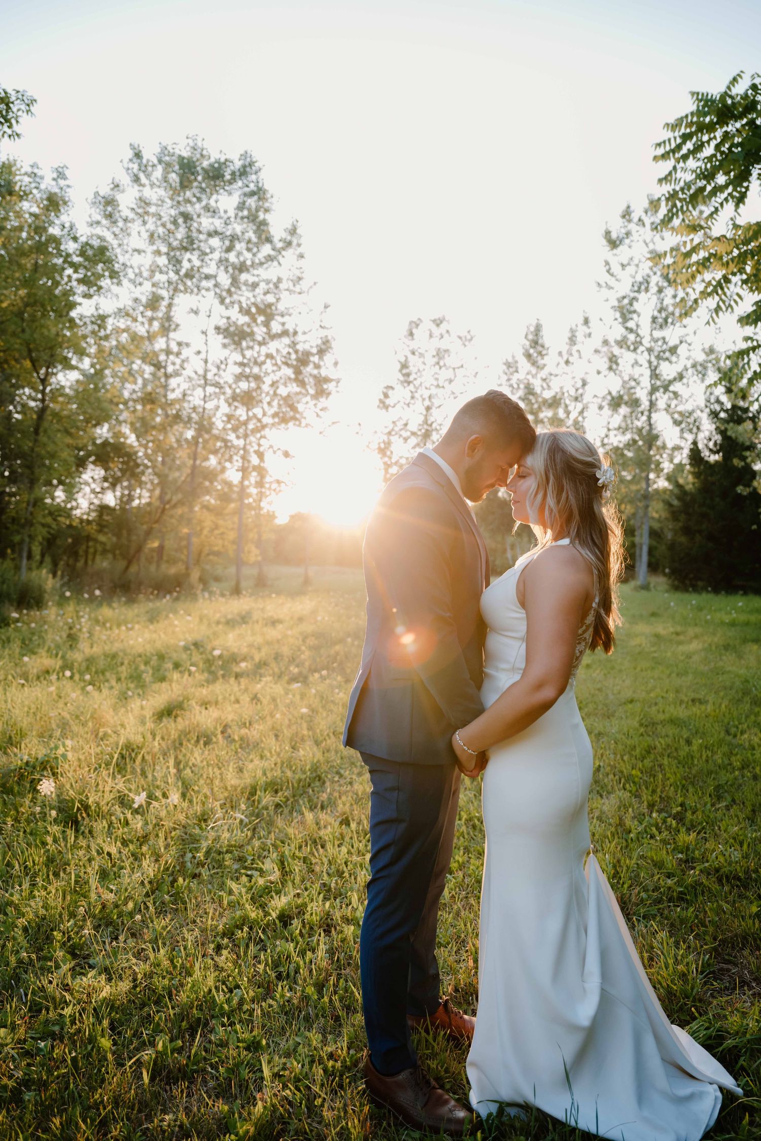 Bride and groom share a romantic moment in a sunlit meadow surrounded by trees at golden hour.