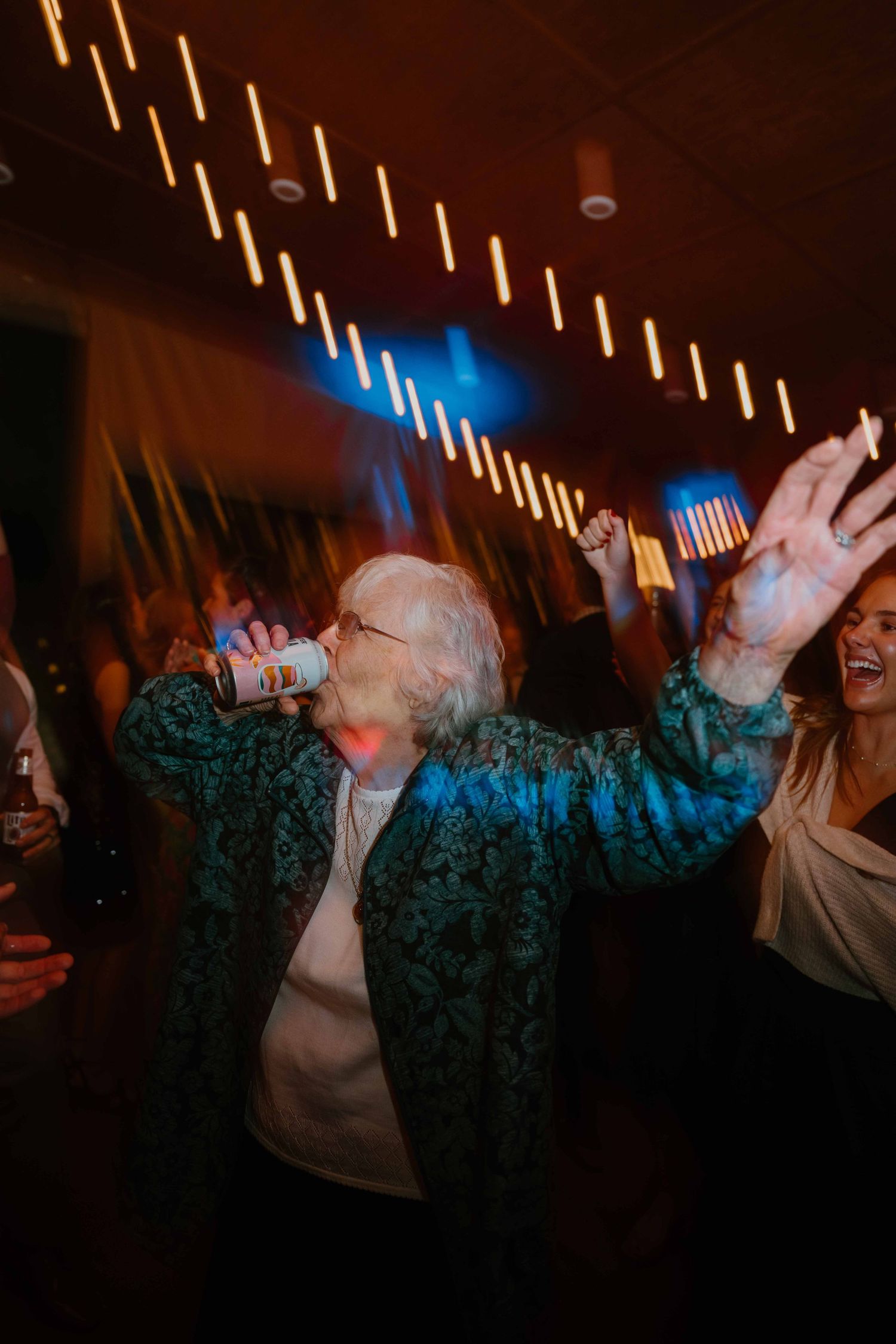 Elderly woman dancing with arms raised while drinking a canned beverage at a wedding reception.
