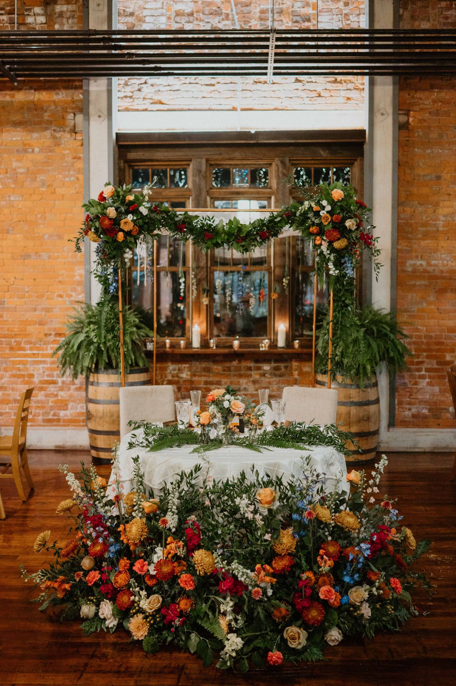 Elegant wedding sweetheart table adorned with colorful floral arch and lush floor arrangements in rustic brick venue.