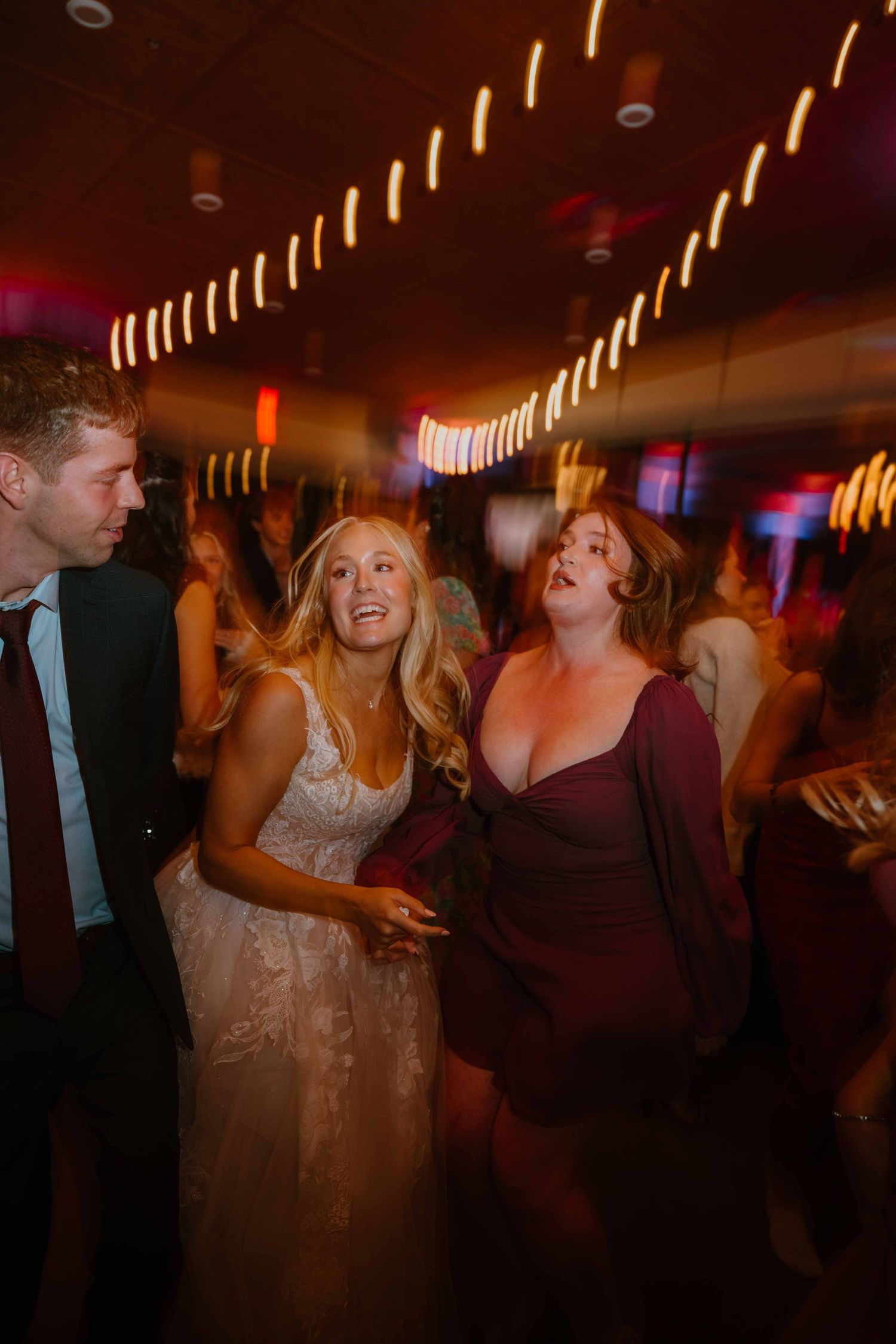 Bride in lace gown dancing joyfully with guests under colorful lights at wedding reception.