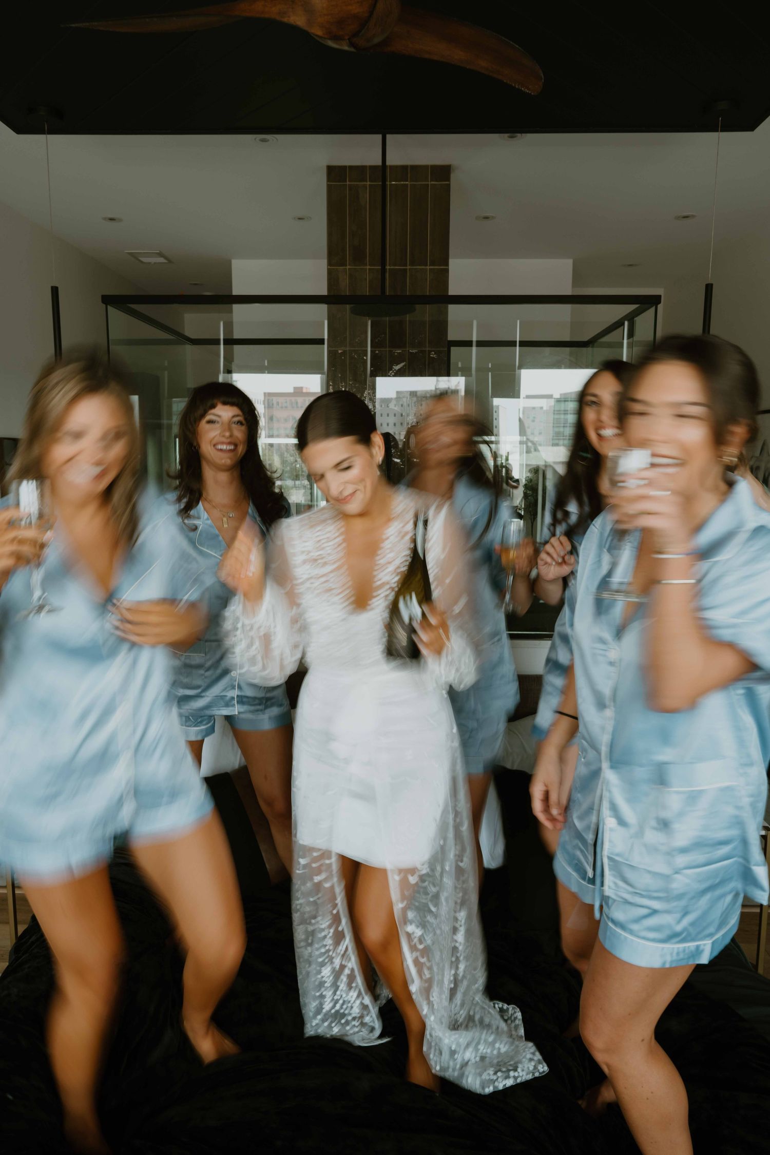 Bride in white lace robe dances with bridesmaids in blue satin pajamas during getting ready.