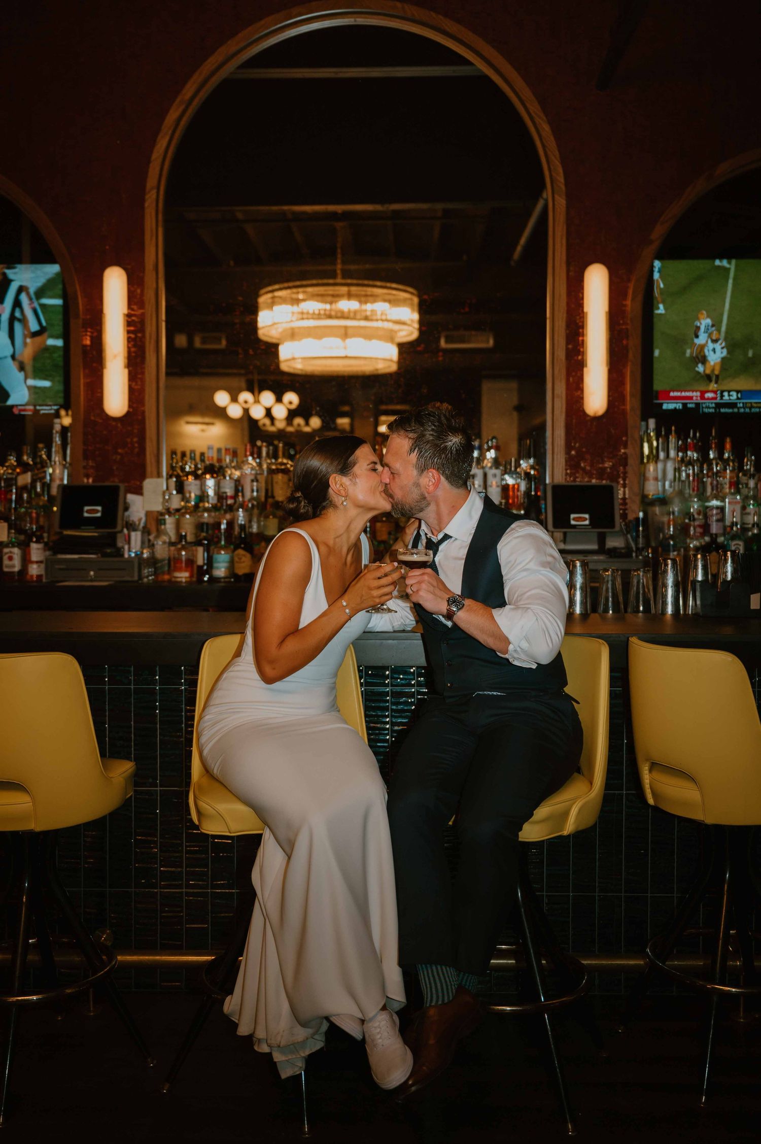 Bride and groom share romantic kiss while seated at elegant bar with yellow stools during wedding reception.