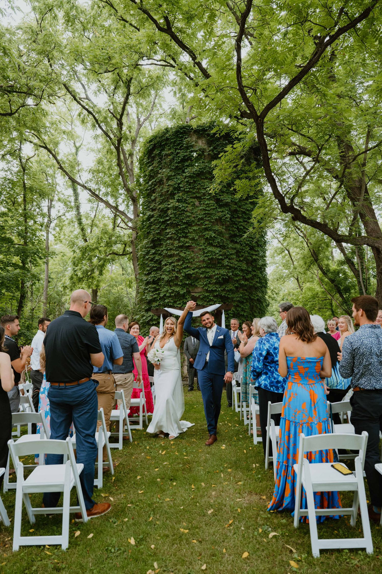 Newlyweds walk joyfully down outdoor aisle surrounded by guests after garden wedding ceremony.