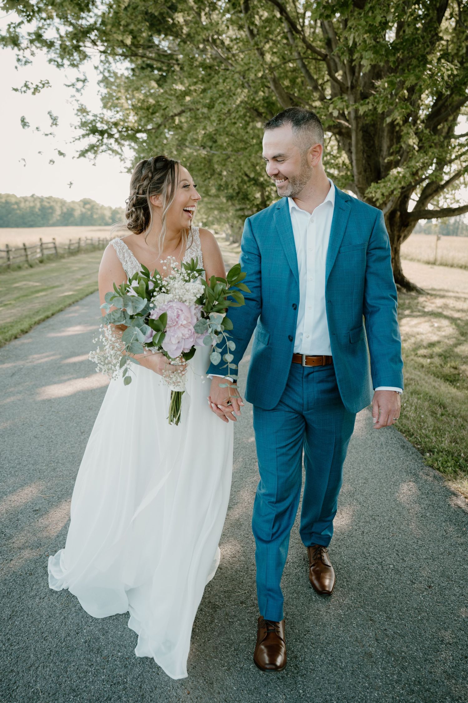 Laughing bride and groom walk hand in hand down tree-lined path with floral bouquet.
