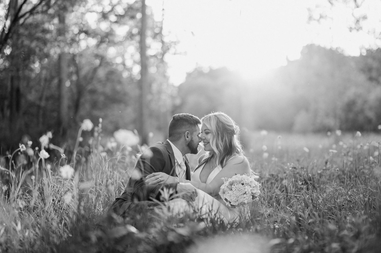Black and white photo of bride and groom sitting close in a wildflower meadow at sunset.