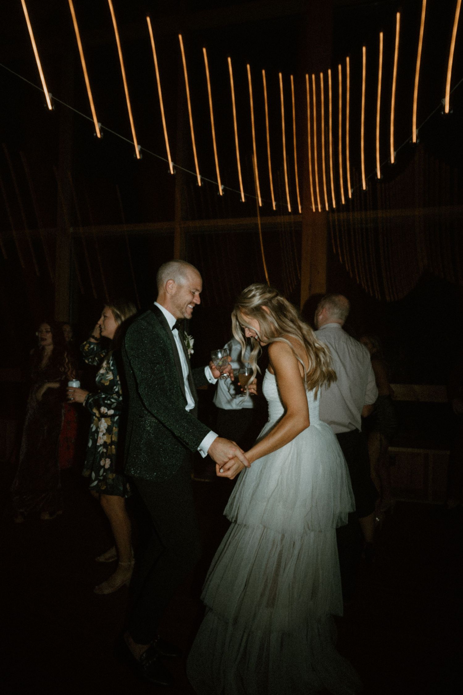 Bride and groom dancing joyfully at wedding reception under warm glowing string lights.
