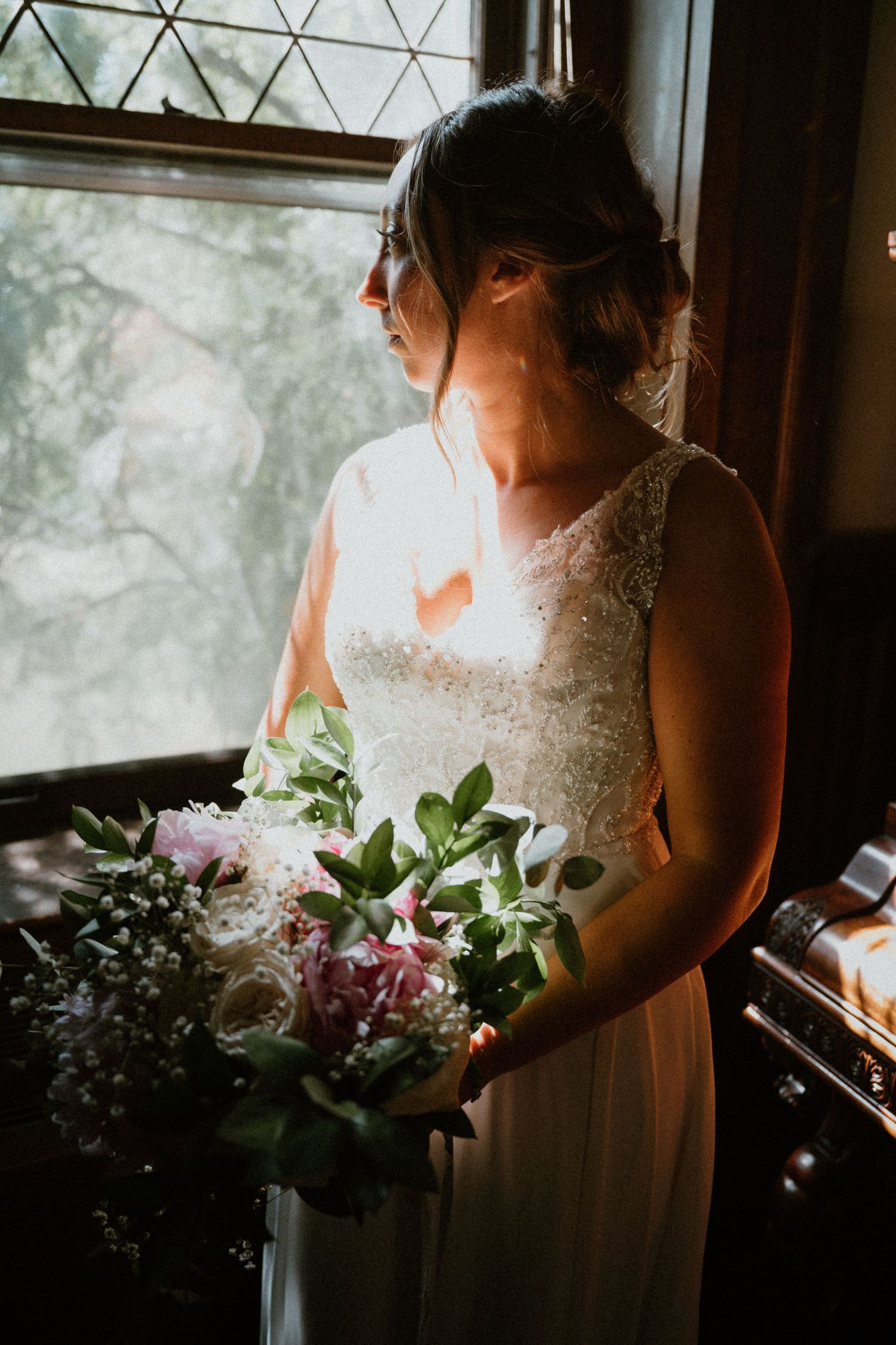 Elegant bride in lace gown holds pink and white bouquet while standing by sunlit window in soft light.