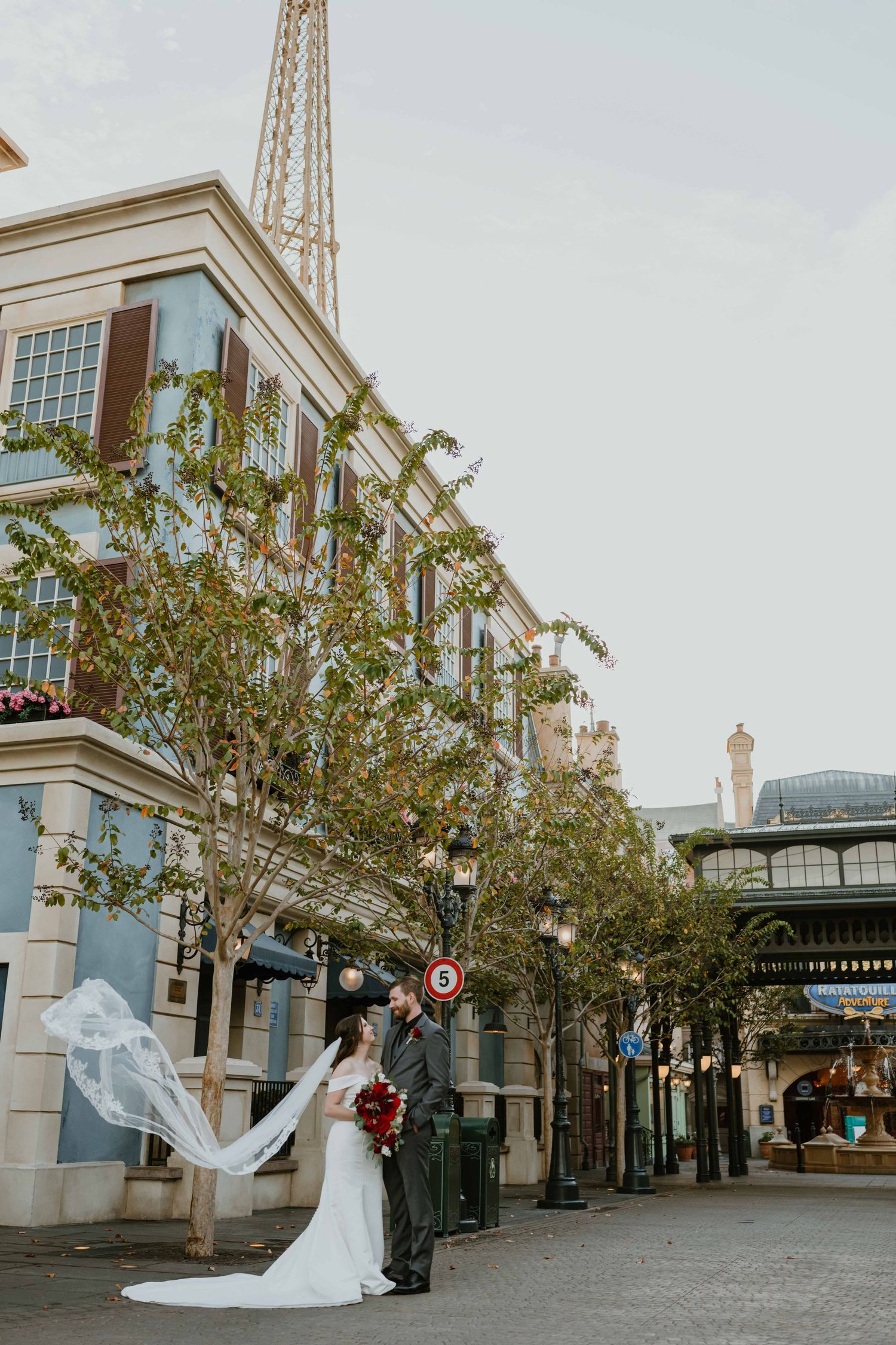 Bride's lace veil billows in the wind as couple embraces on a European-style street at Disney Epcot.