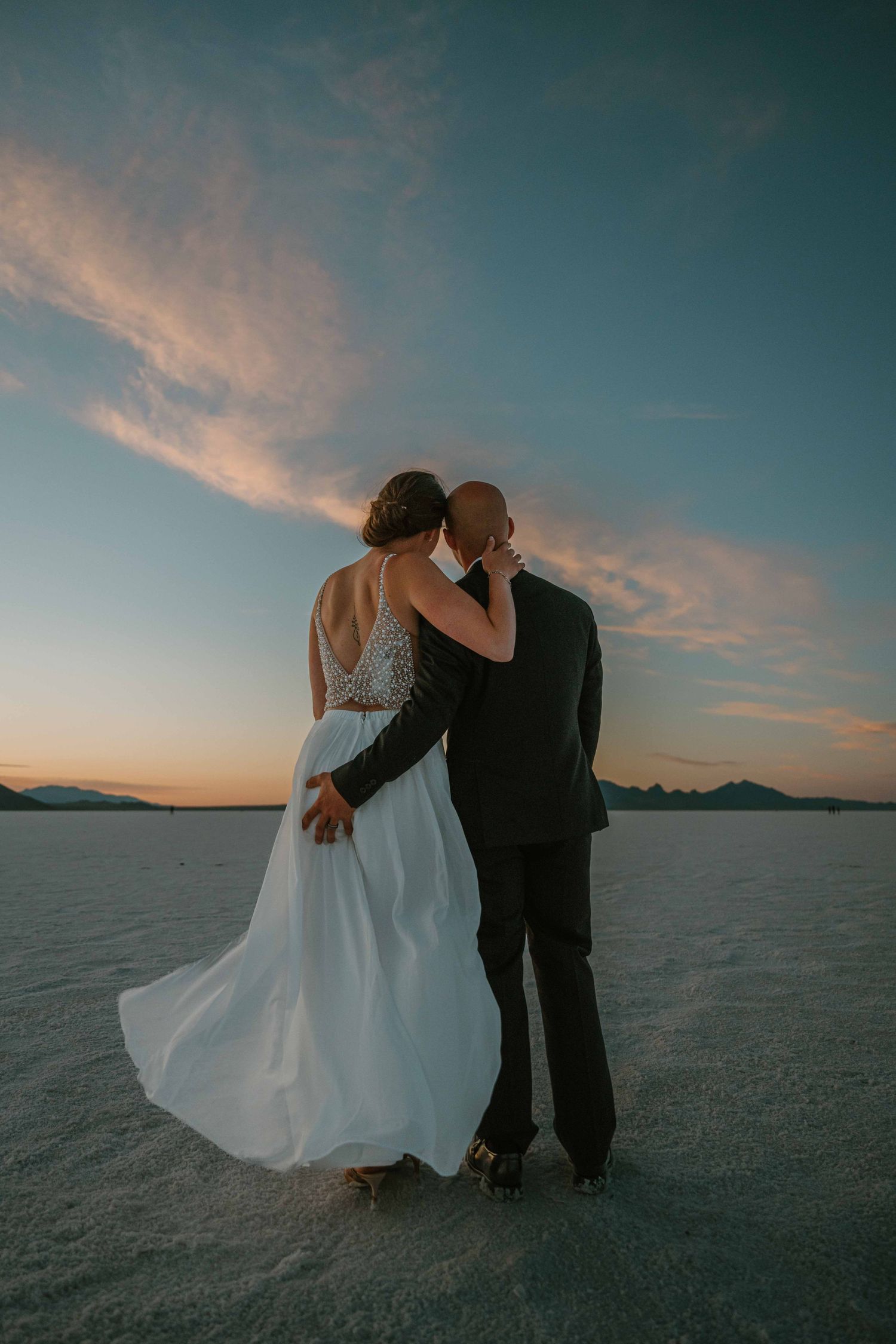 Bride and groom embrace from behind gazing at a stunning sunset sky over the salt flats.