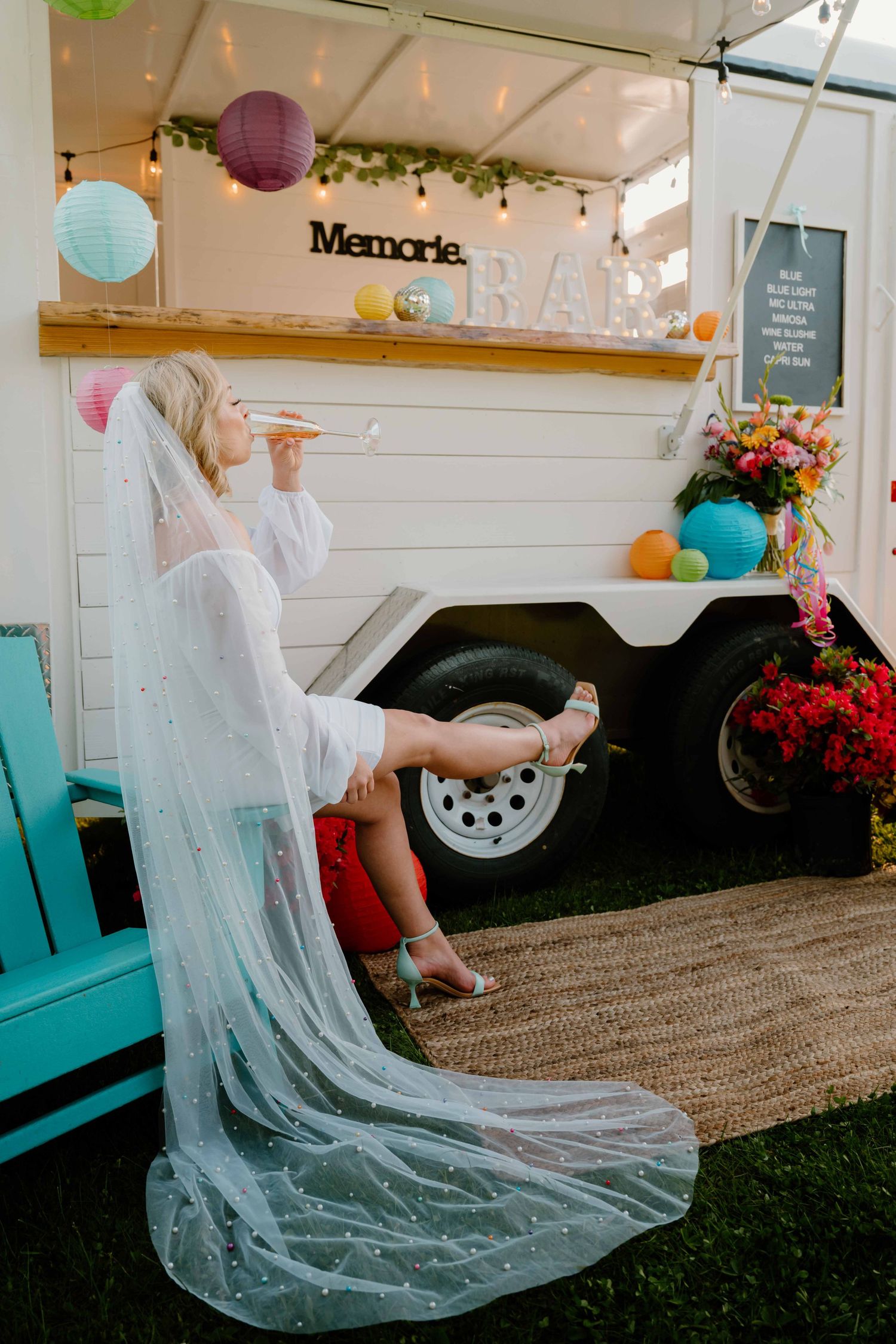 Bride in white gown and veil sits beside a decorated Memories Bar trailer at wedding.