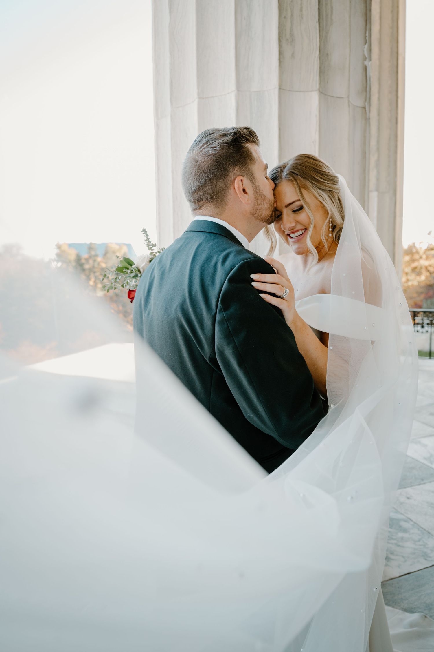 Groom kisses smiling bride on temple as her veil flows between marble columns at wedding venue.
