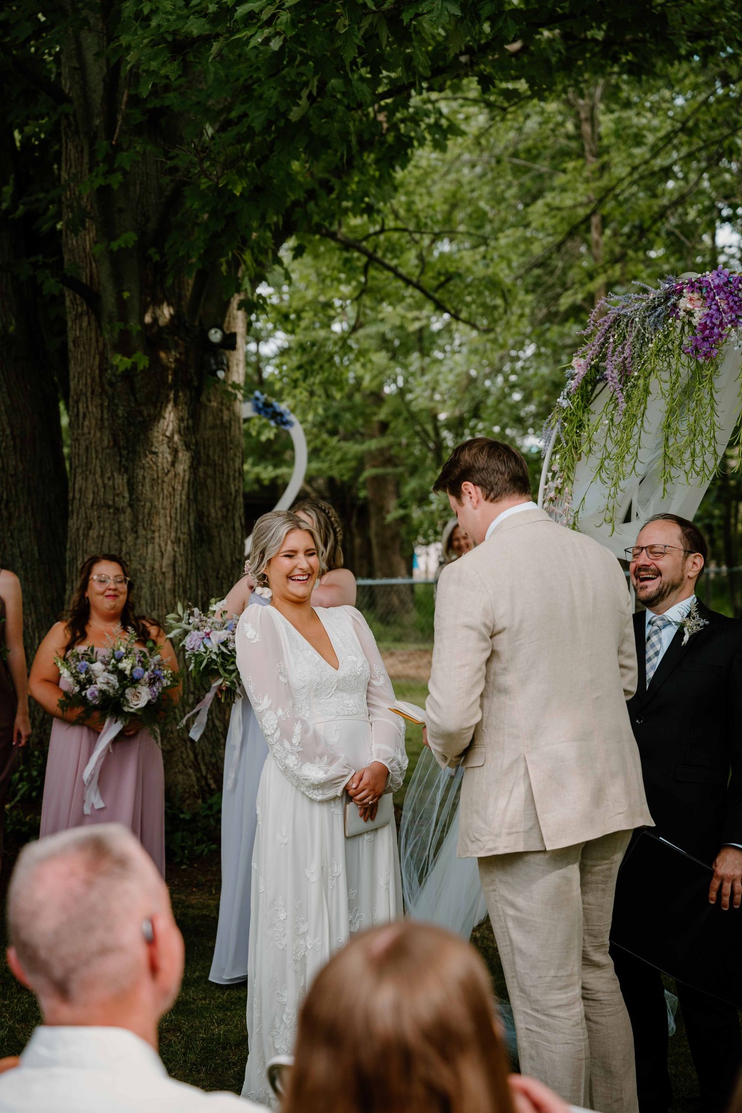 Joyful bride smiles at groom during garden wedding ceremony with purple wisteria arch backdrop.