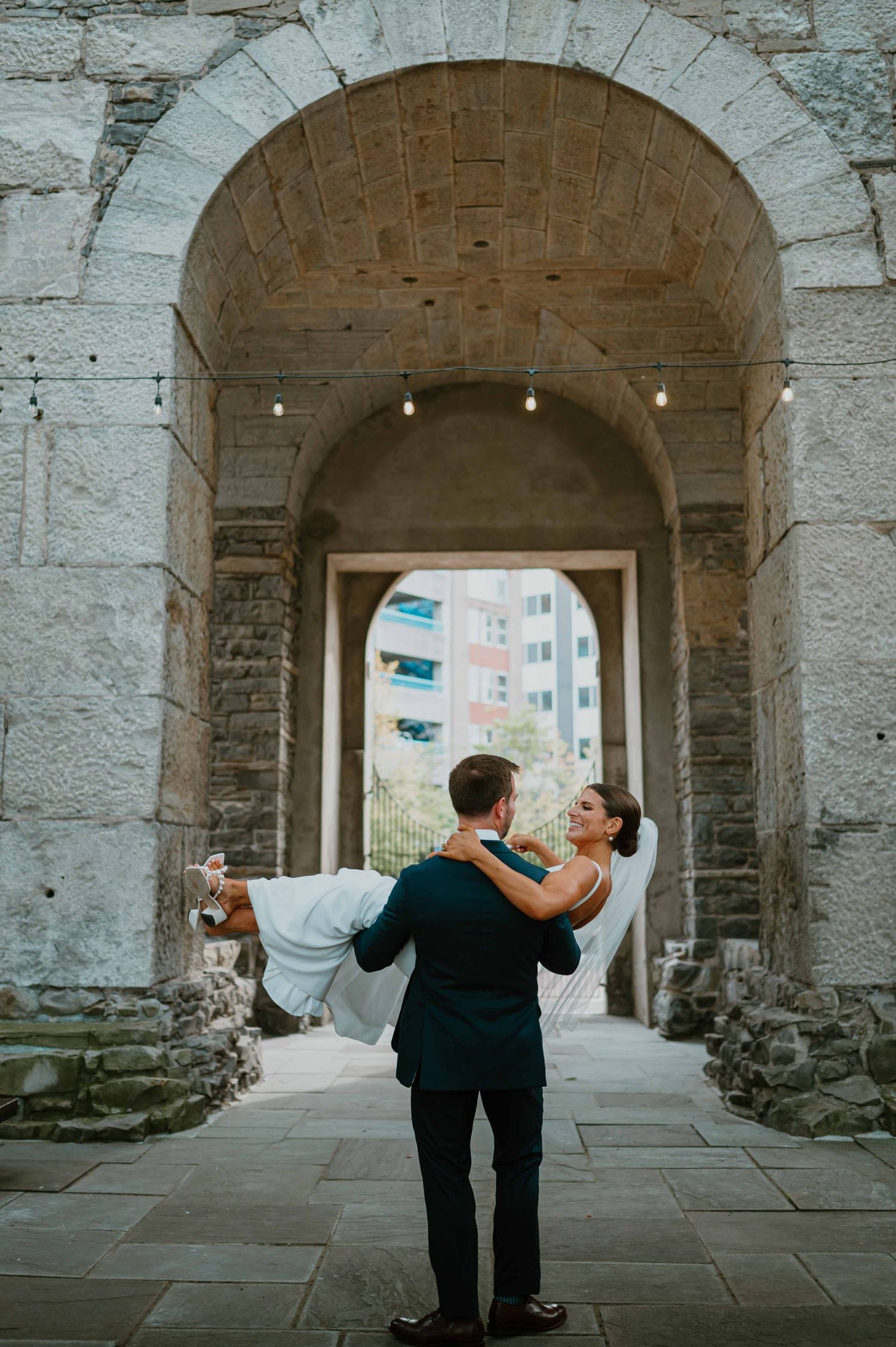 Groom carries bride through a grand stone archway with string lights at an elegant outdoor wedding venue.