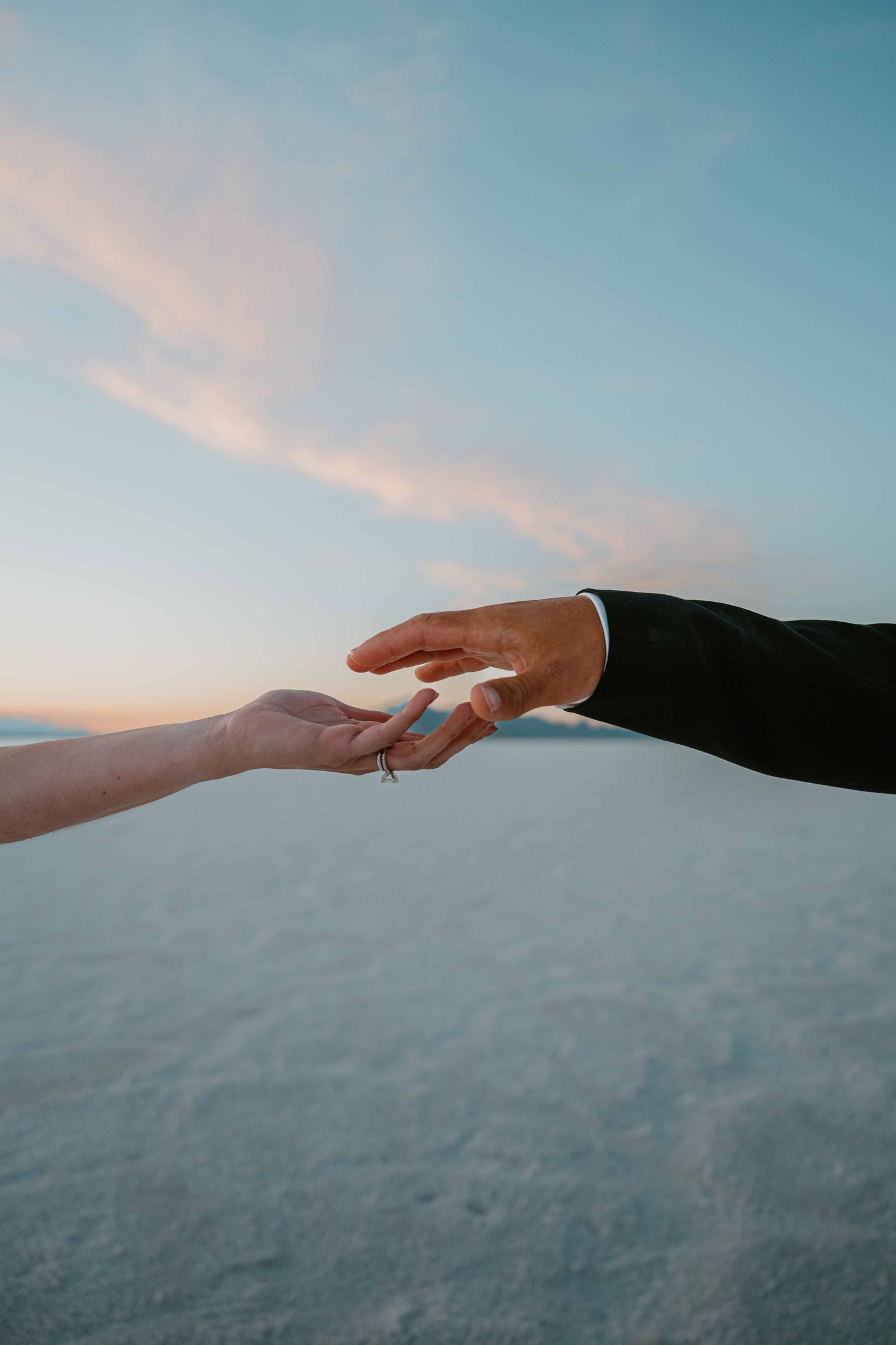 Bride and groom reach toward each other with fingertips nearly touching against a pastel sunset sky.