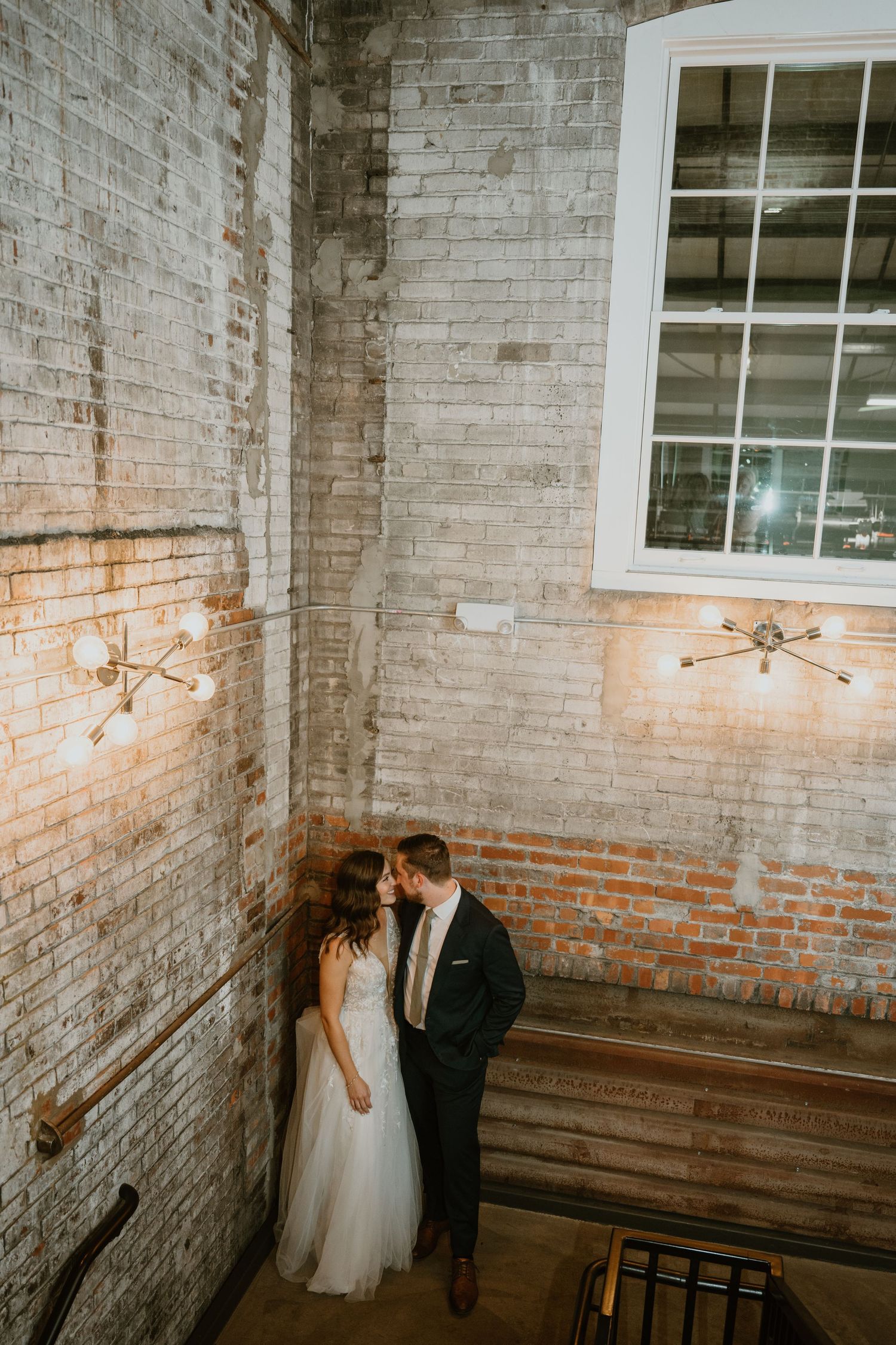Bride and groom share romantic moment against exposed brick wall with vintage sconce lighting.