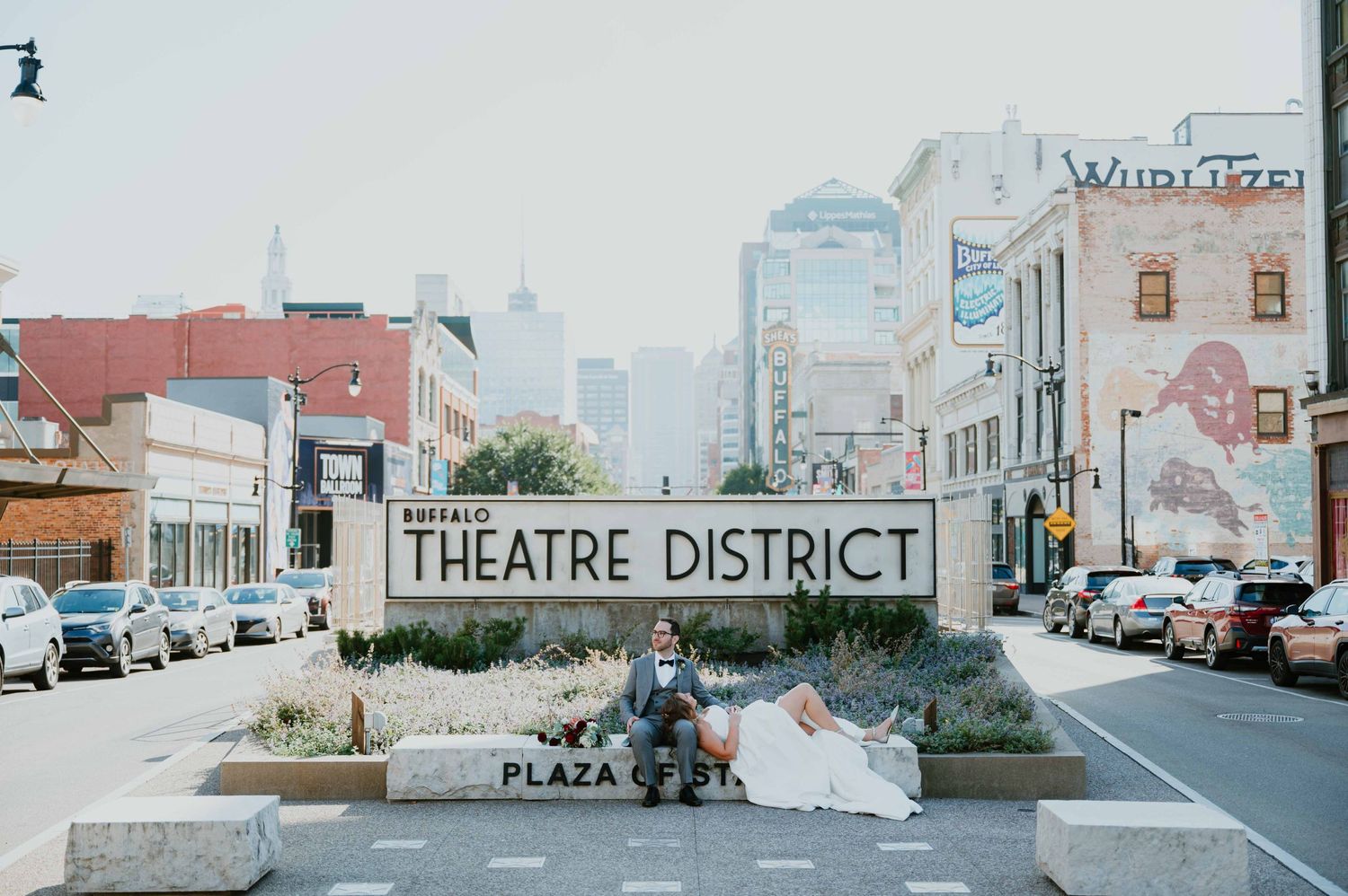 Bride and groom sitting at Buffalo Theatre District plaza sign in urban street setting.
