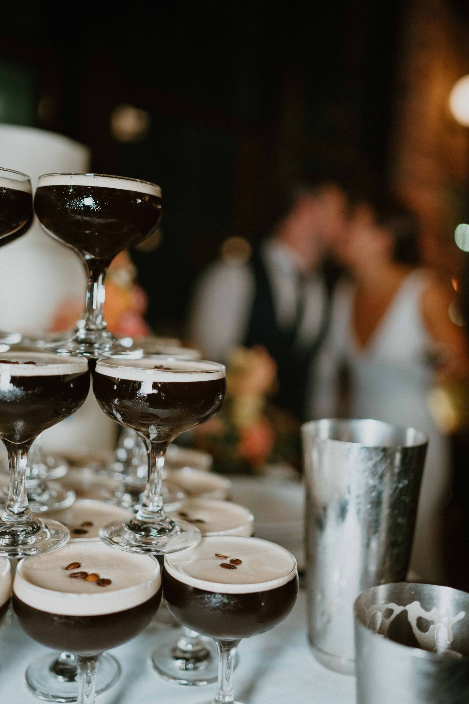 Tower of espresso martini cocktails stacked in coupe glasses with blurred kissing couple in background.