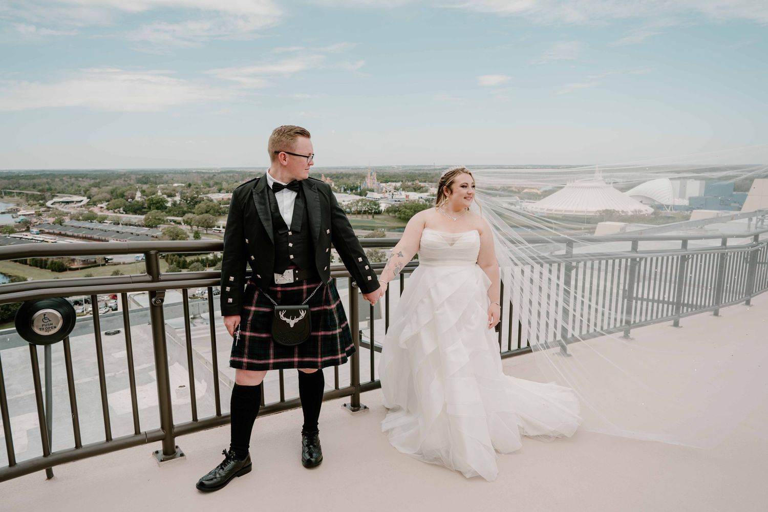 Bride and kilted groom pose on rooftop terrace with panoramic city views on wedding day.