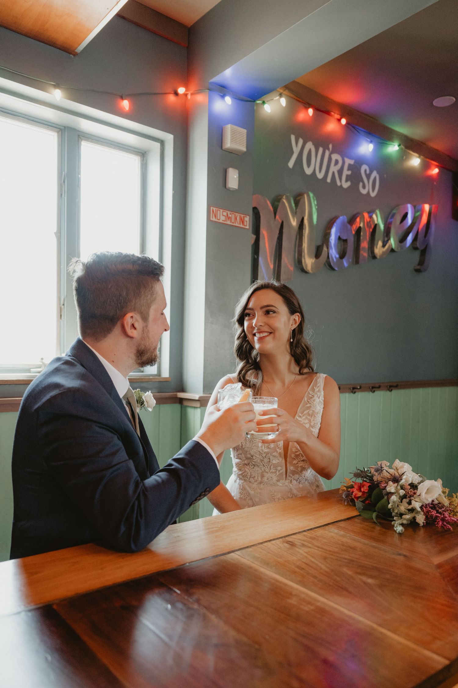 Bride and groom toasting drinks at a bar with colorful lights and fun neon sign backdrop.