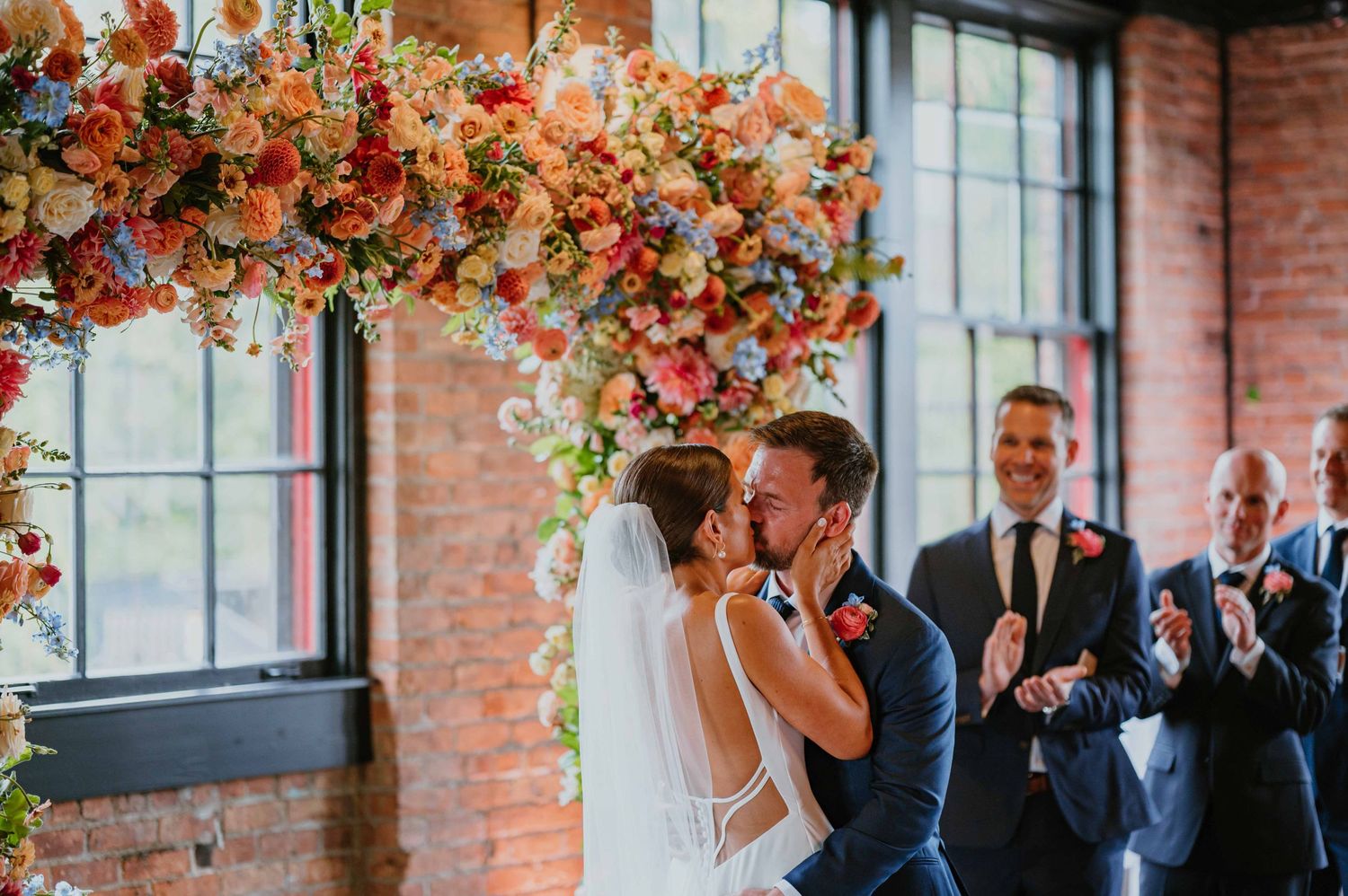 Bride and groom share first kiss under stunning colorful floral arch at indoor wedding ceremony with brick walls.