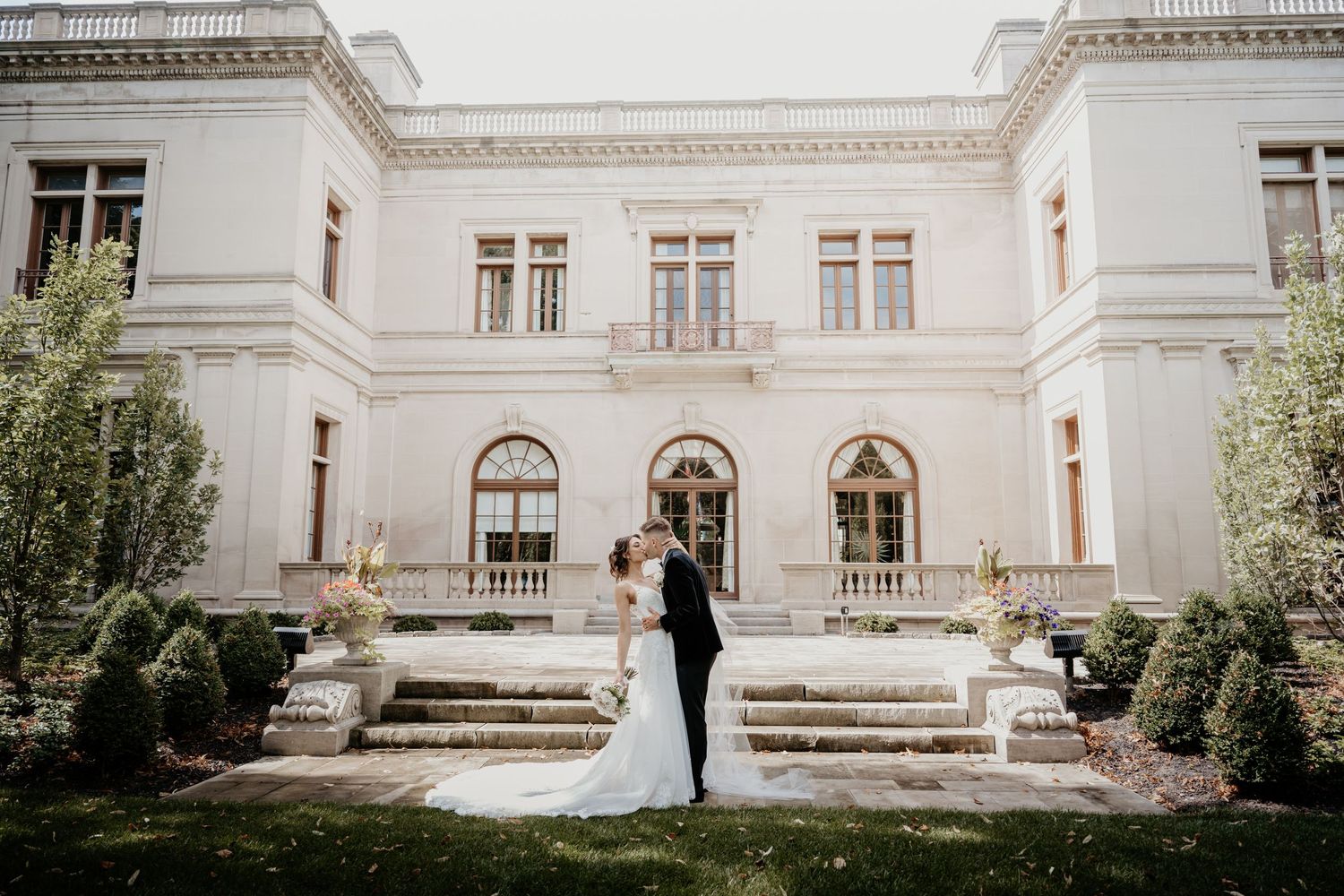 Bride and groom standing before grand white neoclassical estate with manicured gardens.