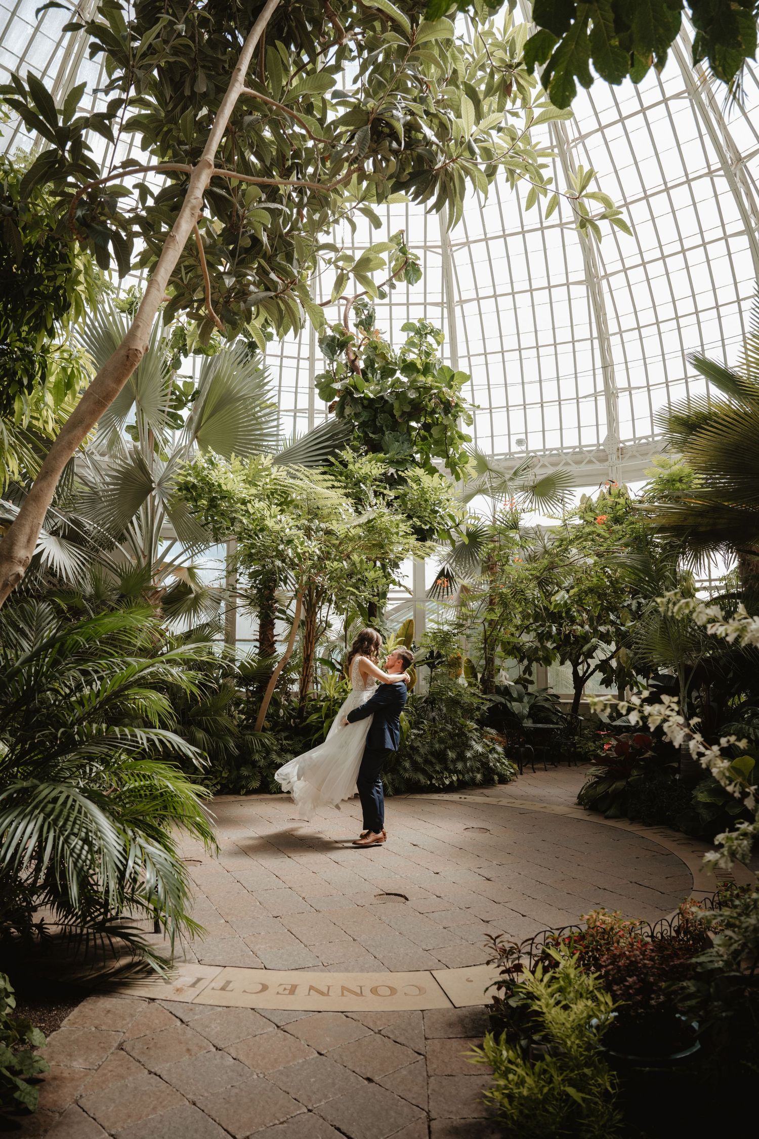 Groom lifting bride in flowing white gown inside a grand botanical conservatory with glass ceiling.