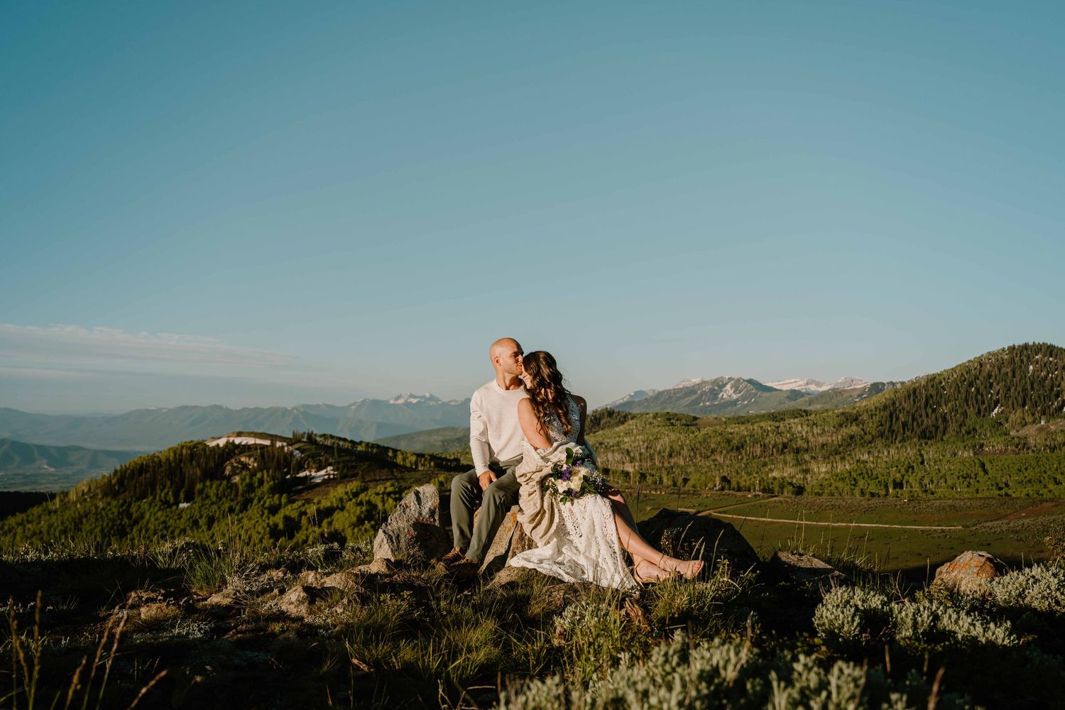 Couple sits closely on rocky hillside with sweeping mountain valley views under clear blue sky.