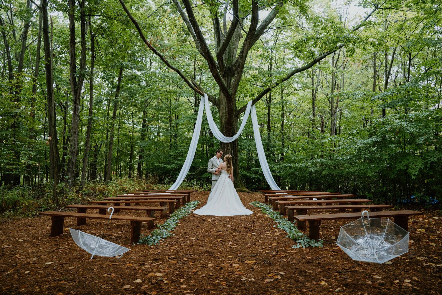 Bride and groom embrace at forest wedding altar with white draping and wooden benches.