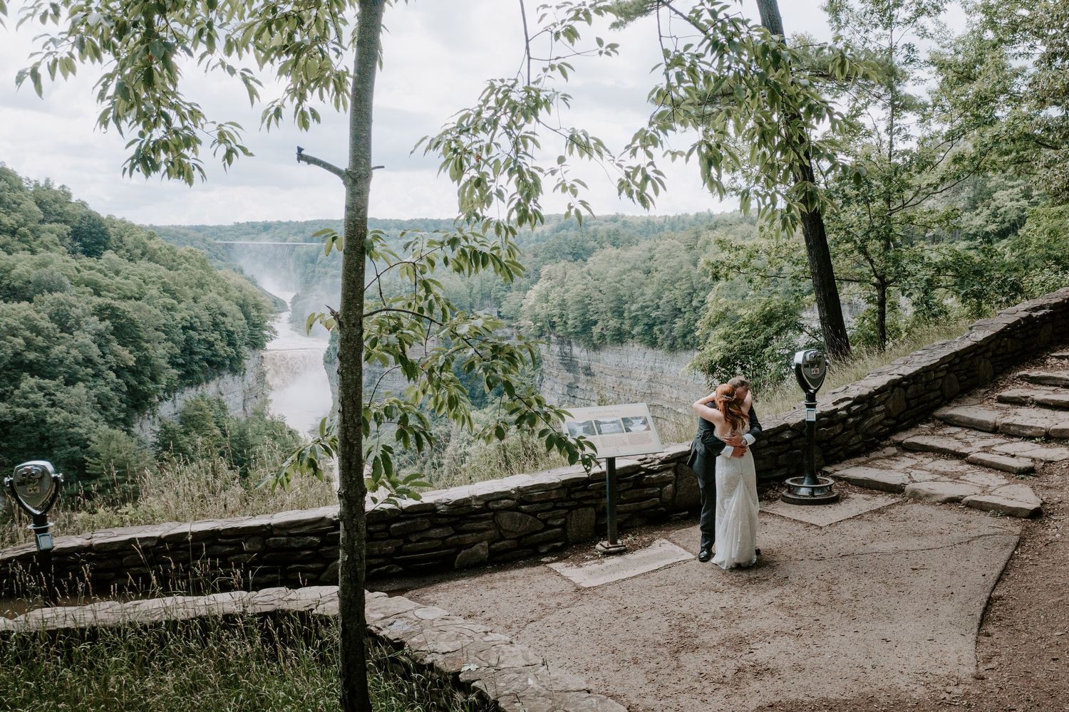 Bride and groom embracing on a stone overlook with a scenic river valley view behind them.
