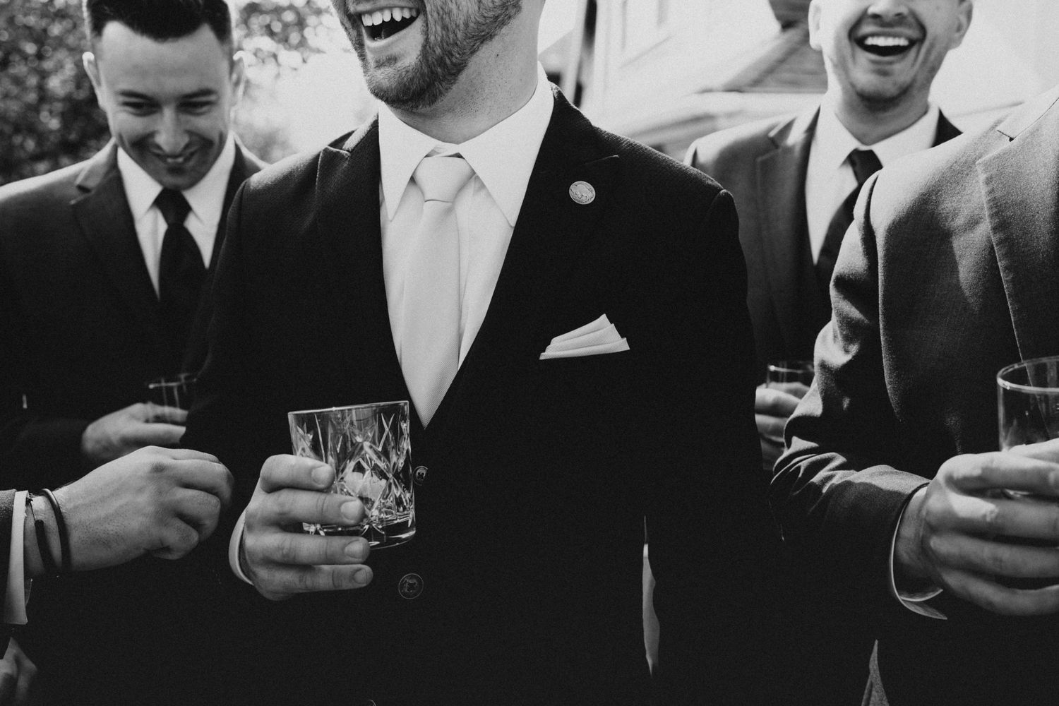 Black and white photo of grinning groomsmen in suits holding whiskey glasses at a wedding.